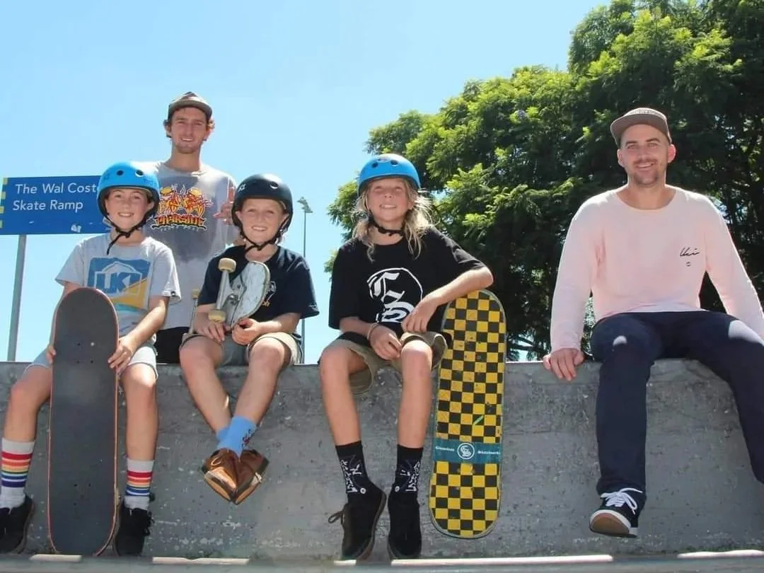 A group of five people, including three children and two adults, sitting on a concrete ledge at a skate park. They are wearing helmets and holding skateboards. The background shows a clear blue sky and some green trees.