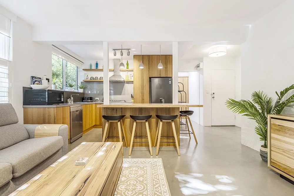 Modern open-plan kitchen and living room with wooden accents, gray sofa, and large potted plant.