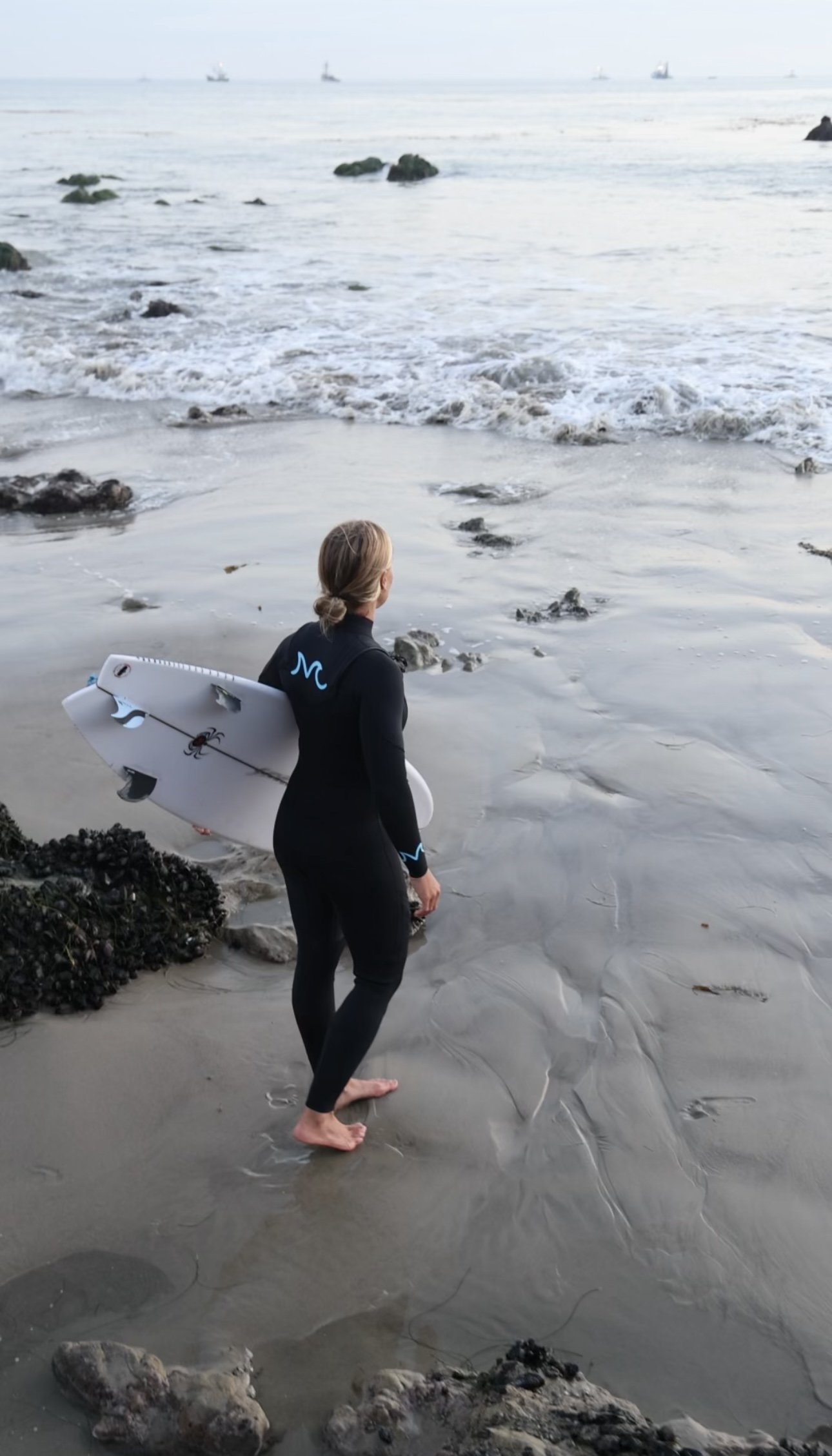 A woman in a black wetsuit with a surfboard standing on a sandy beach near the water's edge, looking out at the ocean.