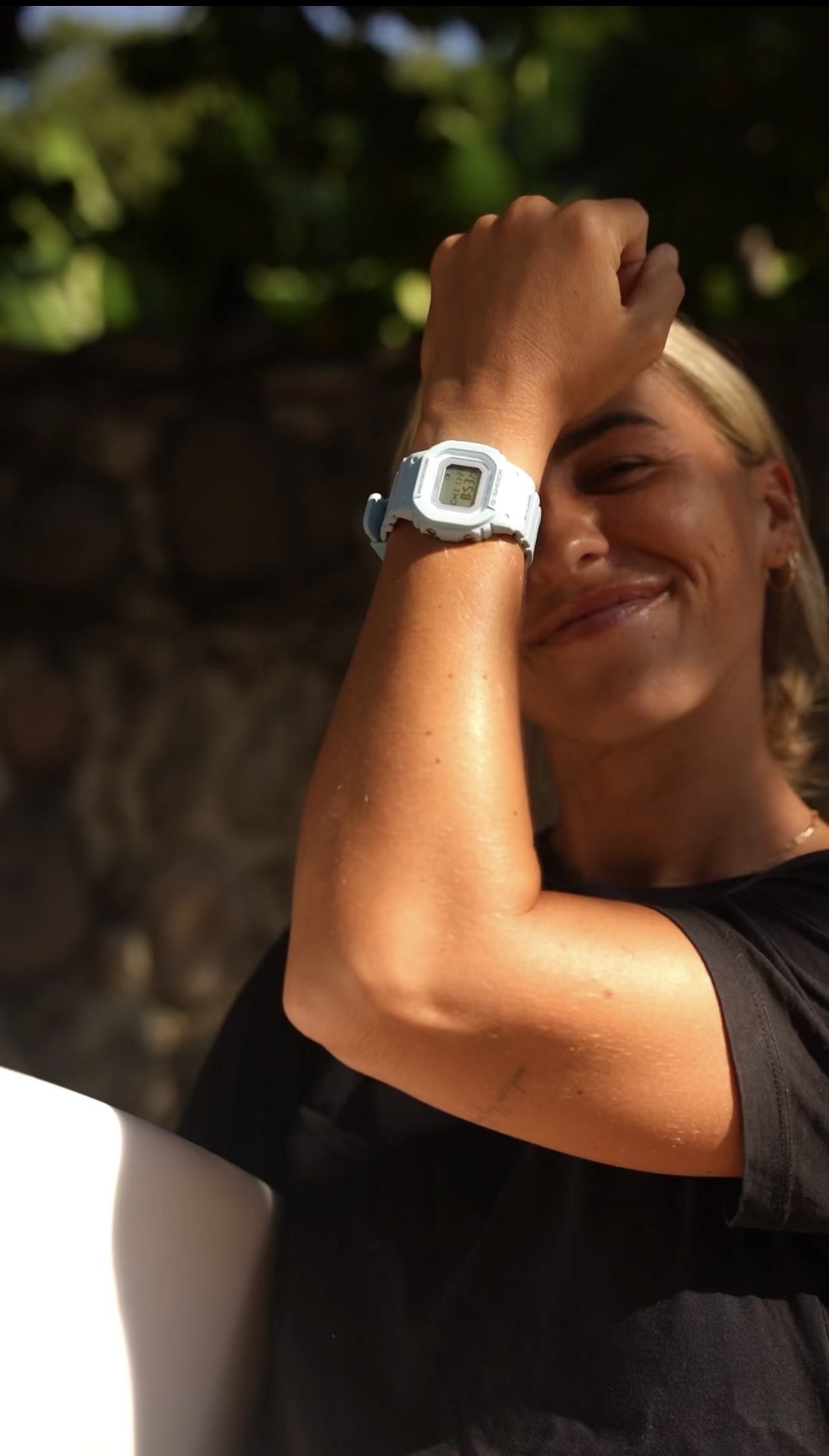 A smiling woman with blond hair, wearing a white digital watch, seated outdoors with a blurred natural background, resting her forehead on her hand.