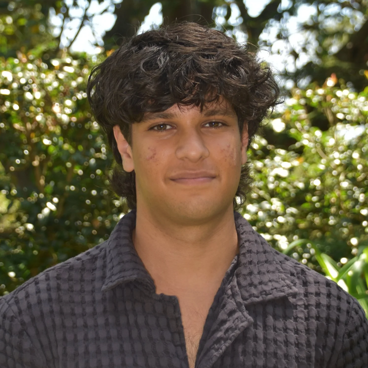 A young man with dark, curly hair and acne on his face, wearing a dark quilted shirt, standing outdoors in front of green foliage.