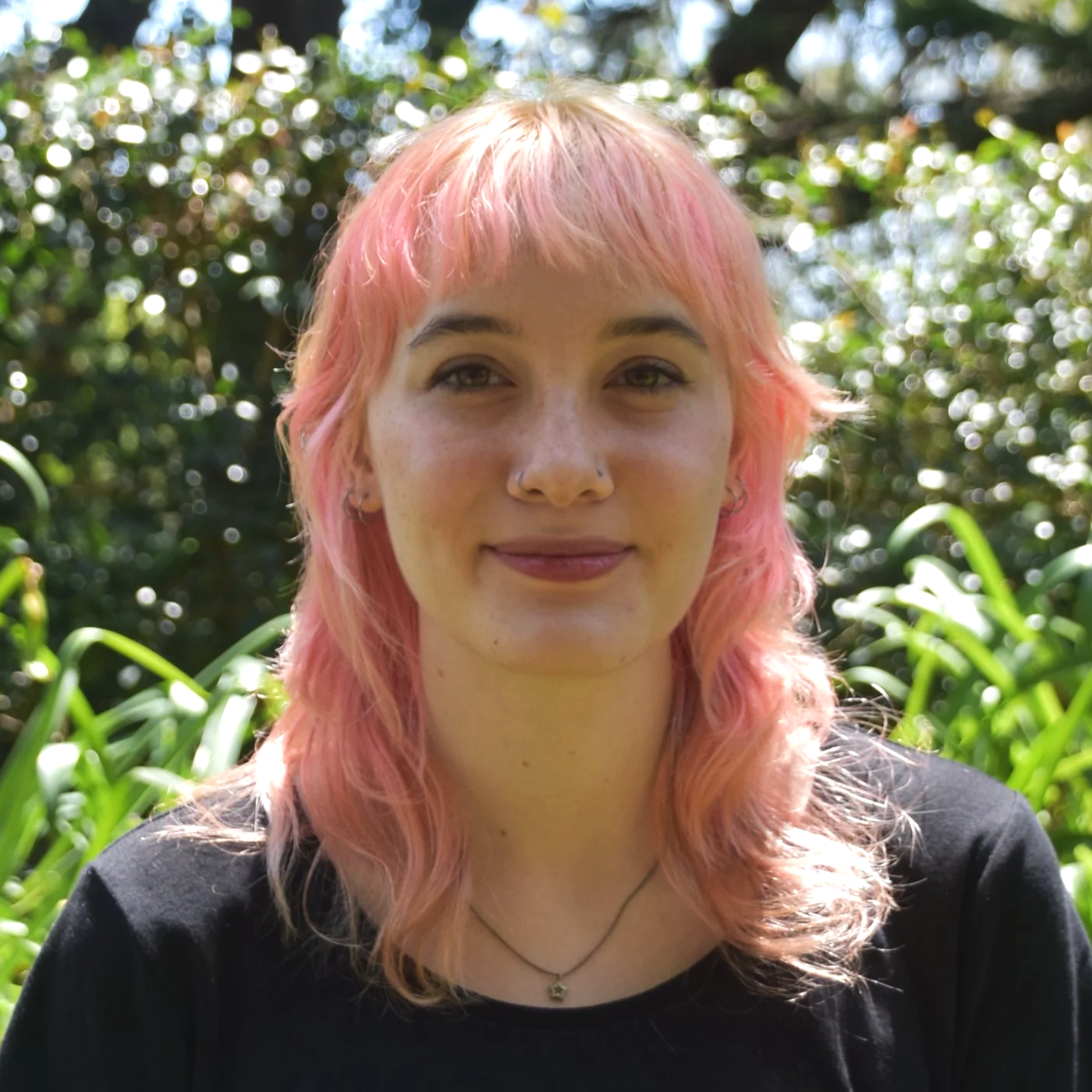 A young woman with pink, shoulder-length hair, wearing a black top and a necklace, standing outdoors in a green, leafy environment with sunlight filtering through the trees.