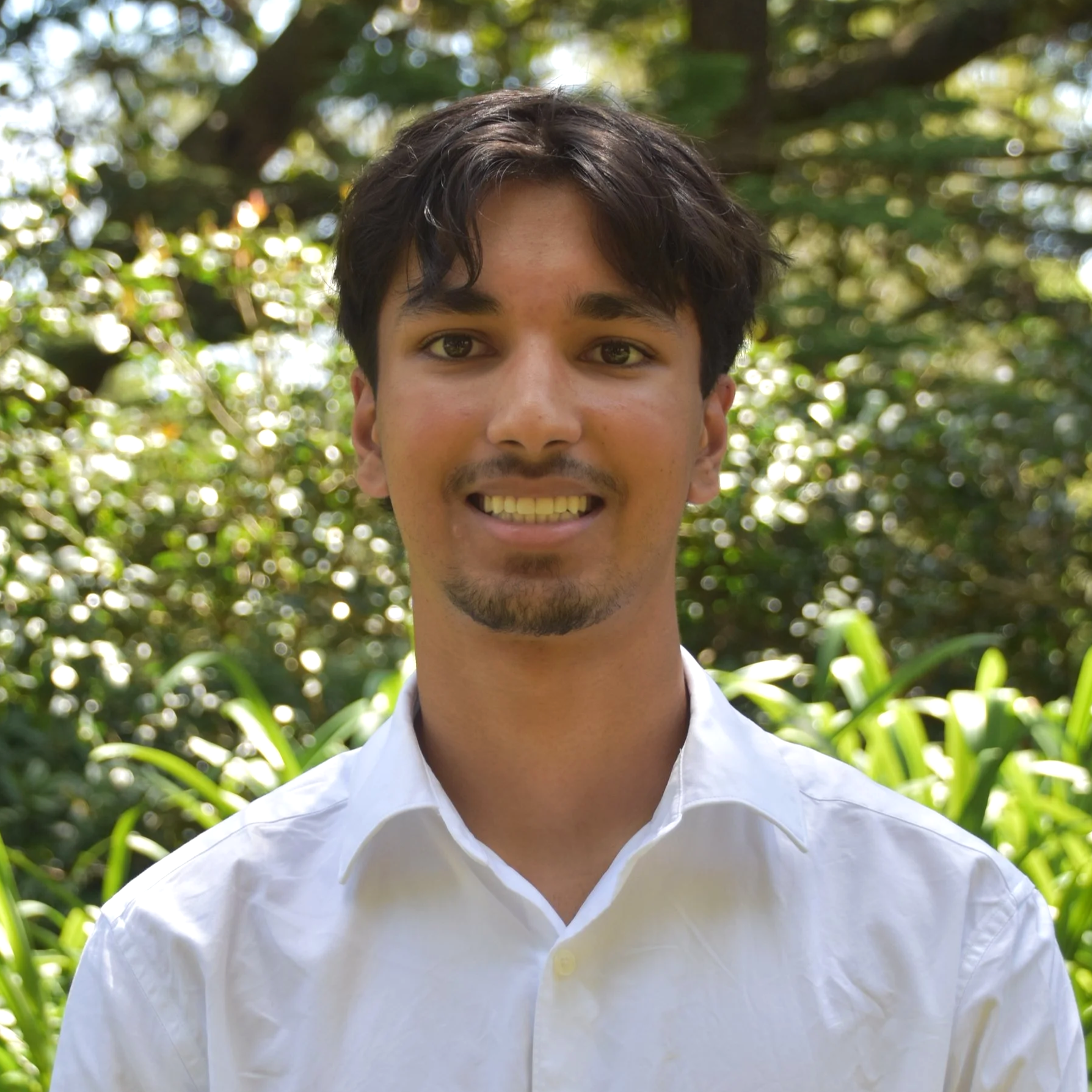 A young man with dark hair and a goatee, wearing a white collared shirt, smiling outdoors with green trees and sunlight in the background.