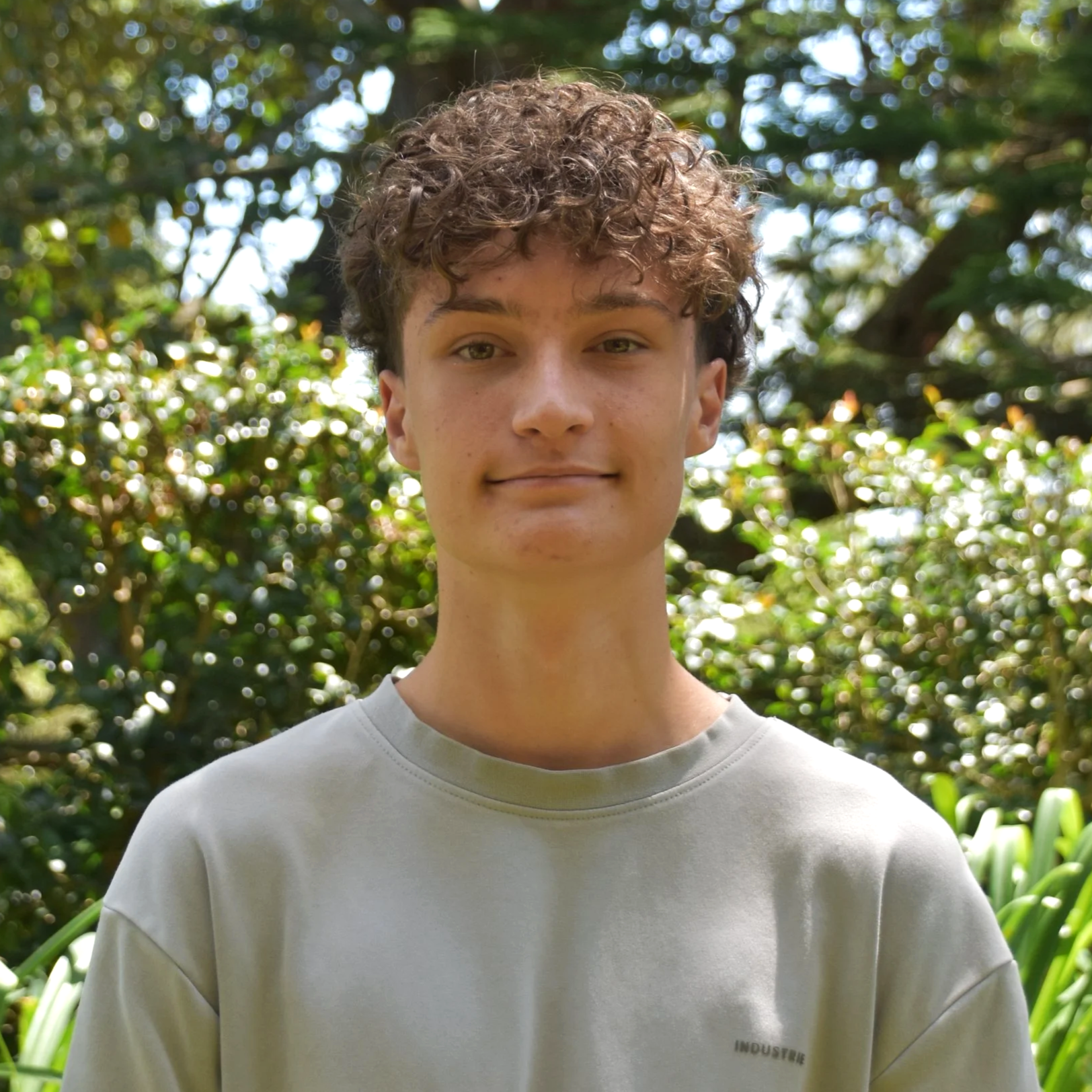 A young man with curly brown hair and freckles stands outdoors in front of lush green trees and plants, wearing a light gray sweatshirt with the word 'INDUSTRIE' on it.