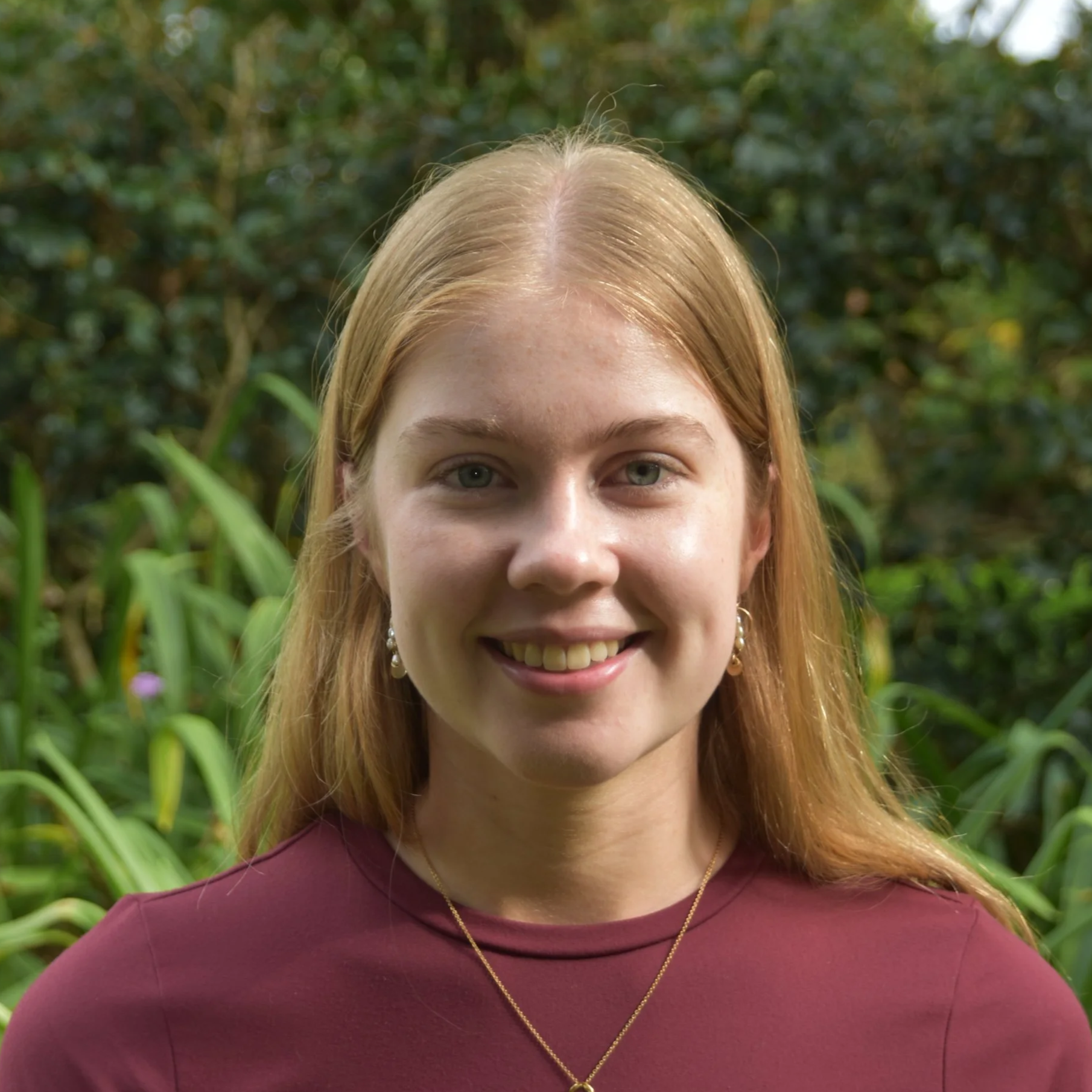 A young woman with red hair smiling outdoors, wearing a maroon top and gold jewelry, with green foliage in the background.