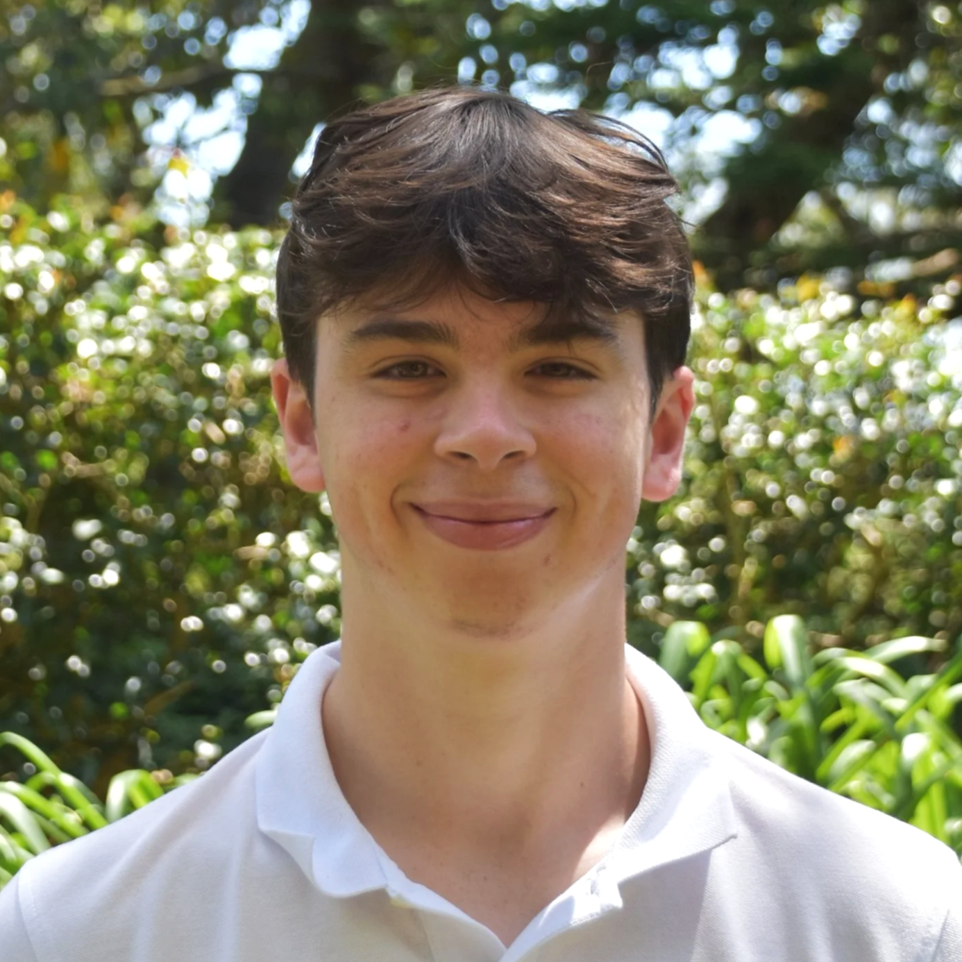 A young man with dark brown hair and fair skin smiling outdoors in a lush green environment.