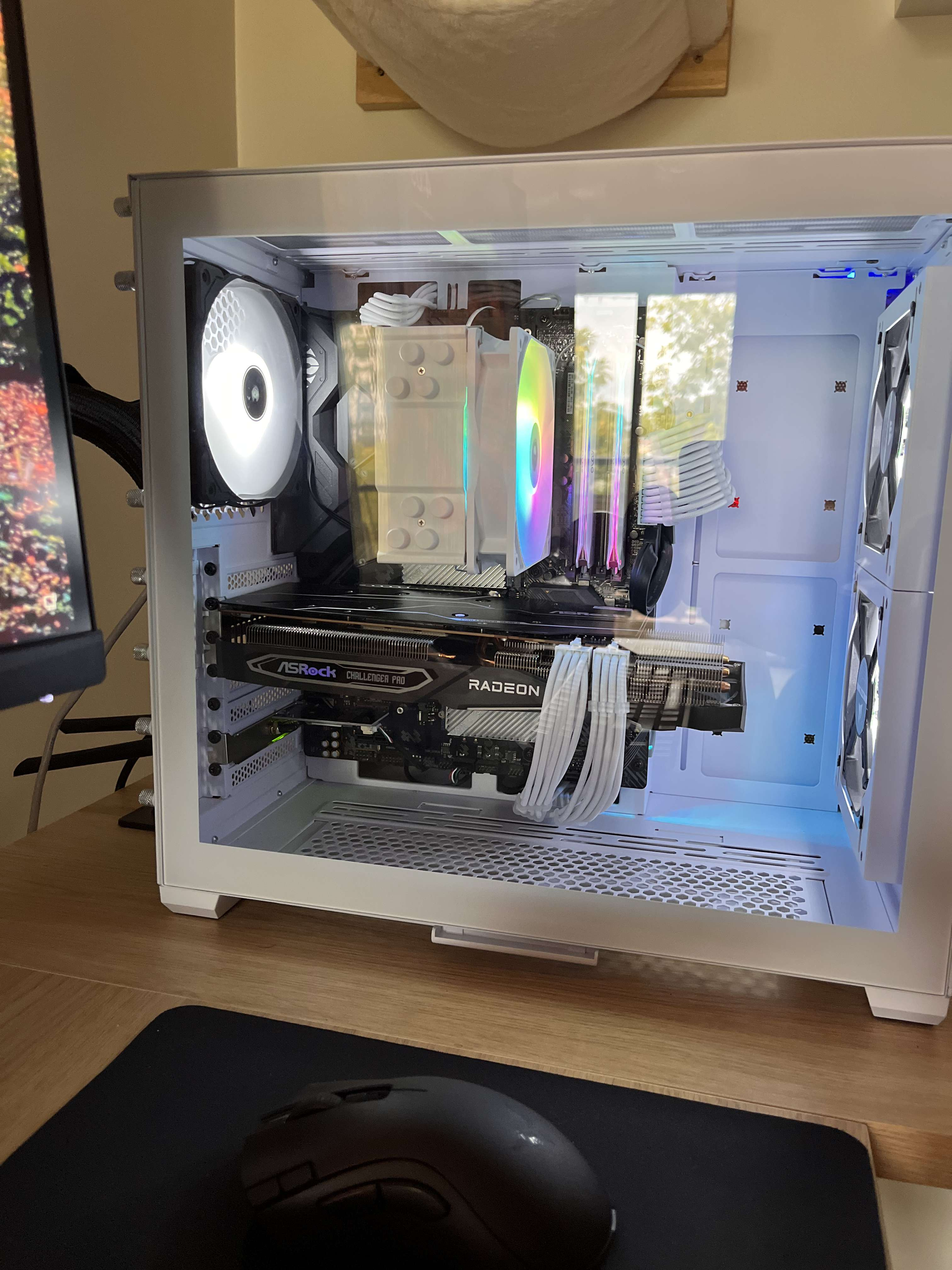 Inside a white computer case with RGB fans, an RGB CPU cooler, a Radeon graphics card, and white cables, placed on a wooden desk with a monitor and mouse nearby.