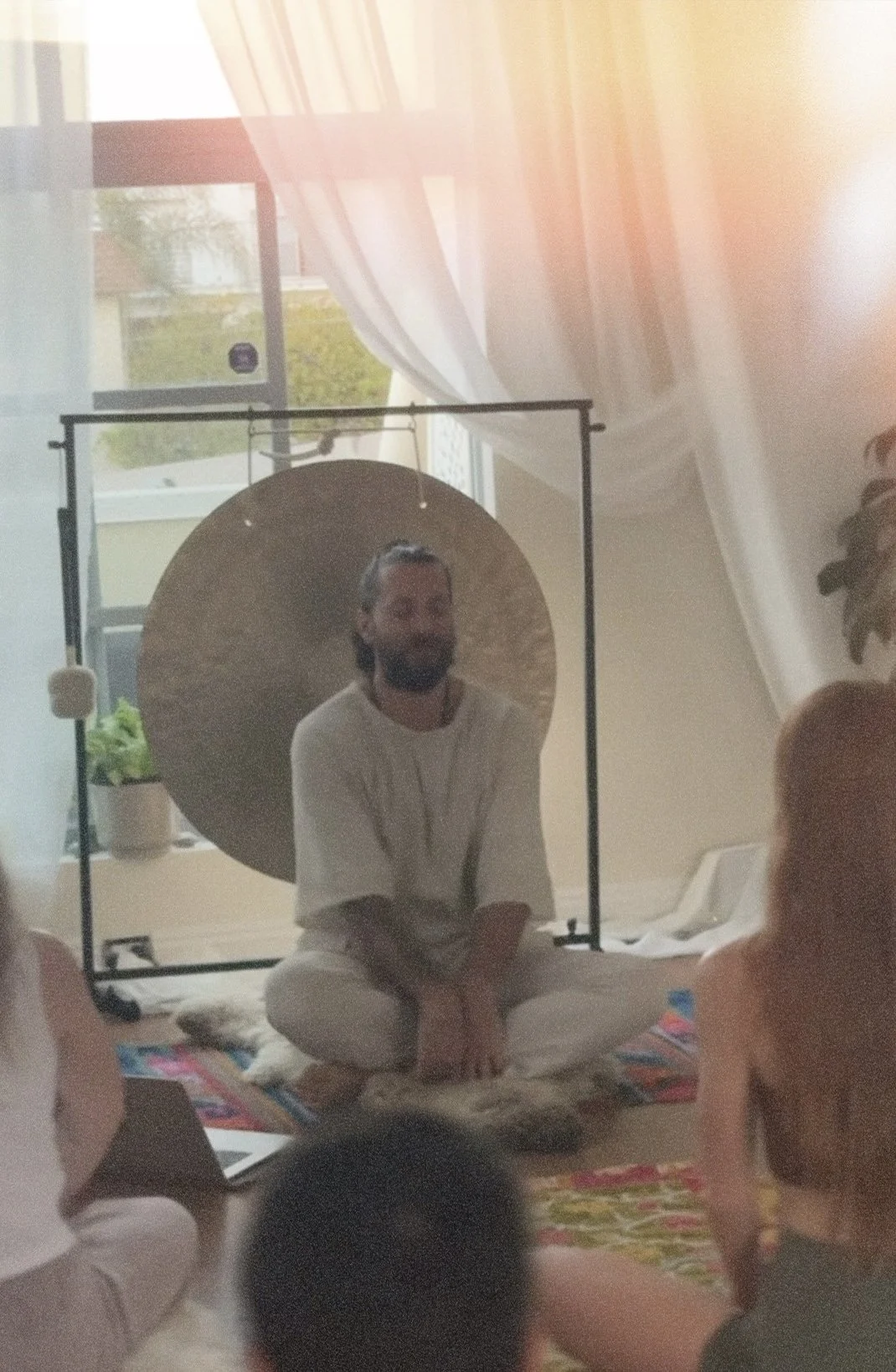 A man with long hair sitting on the floor in front of a group of people, teaching a kundalini yoga class and meditation with a large gong behind him, inside a room with a window and curtains partly open, allowing sunlight to enter.