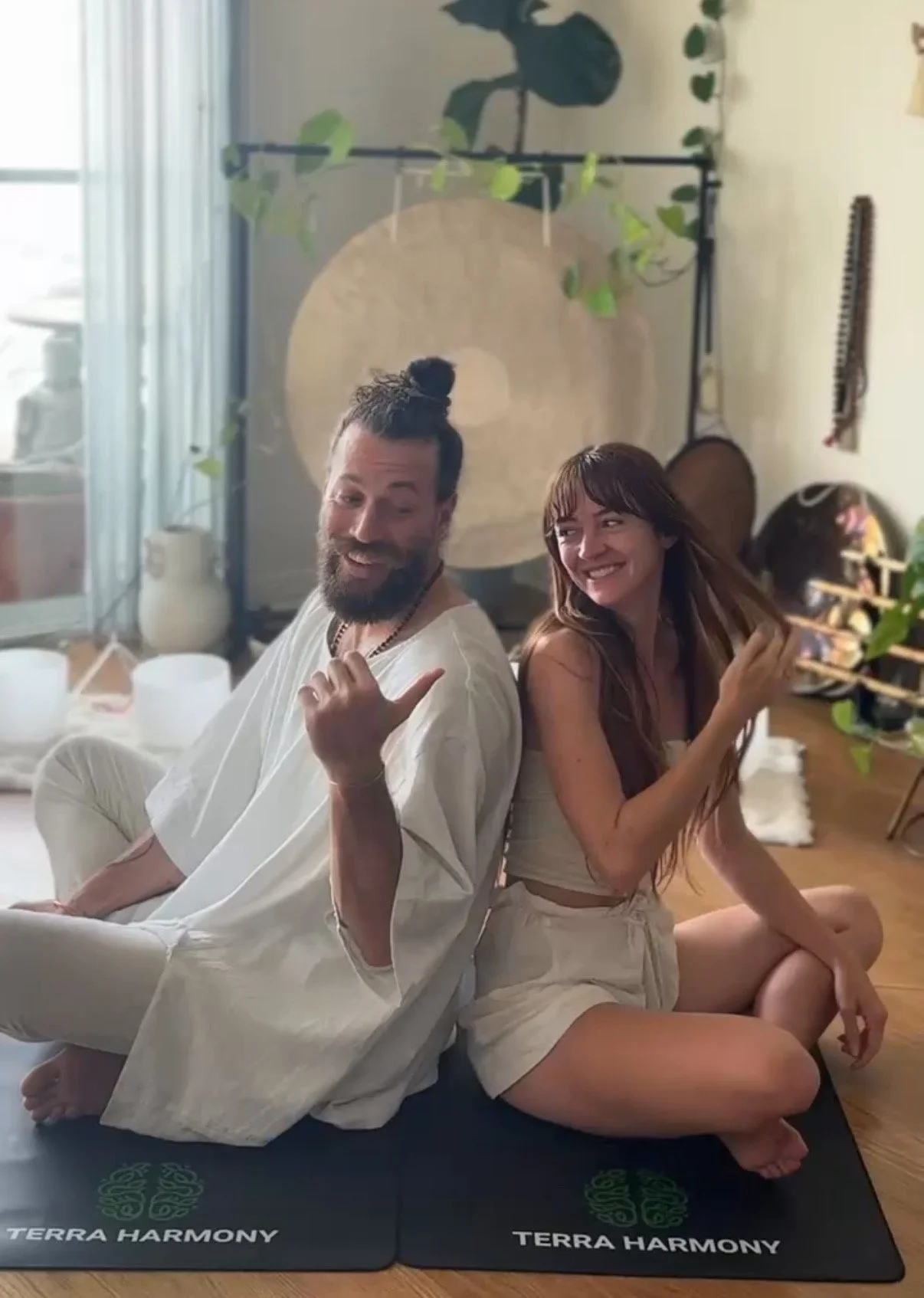 A man with long hair sitting on the floor in front of a group of people, teaching a kundalini yoga class and meditation with a large gong behind him, inside a room with a window and curtains partly open, allowing sunlight to enter.
