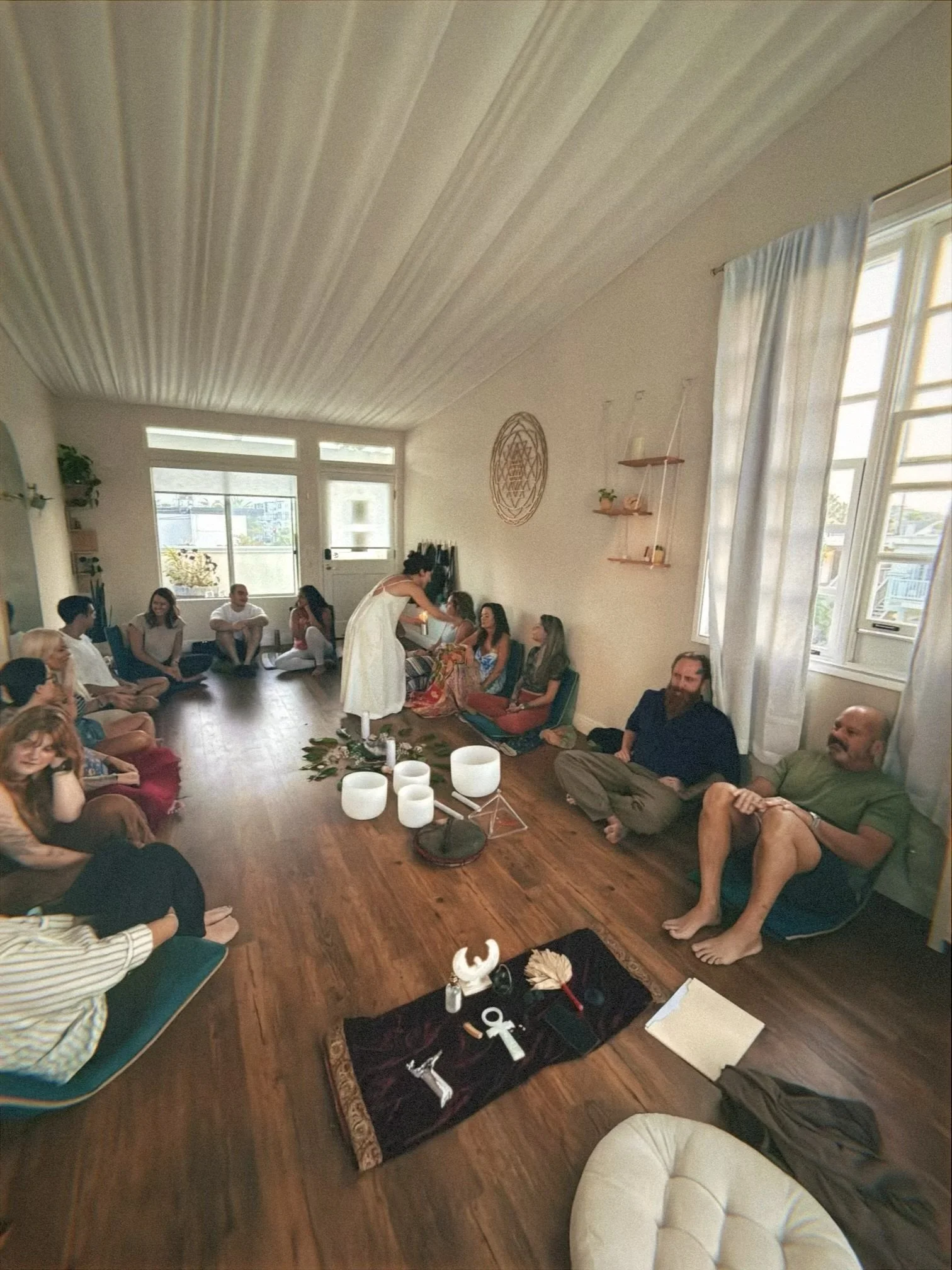 A group of people sitting on the floor in a circle in a cozy, well-lit room with wooden floors and white walls. A woman in a white dress is standing in the center, performing a ceremony with various objects like candles and crystals on a black cloth on the floor in front of her.