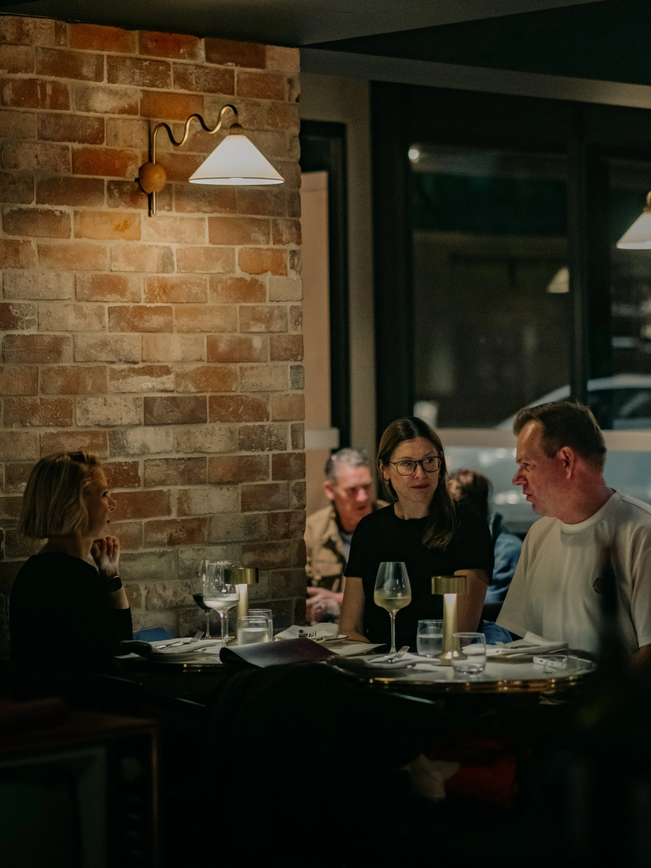 People dining together at Annabel's Wine Bar, sitting around a table with wine glasses and candles, inside a cosy space with brick walls and large windows.