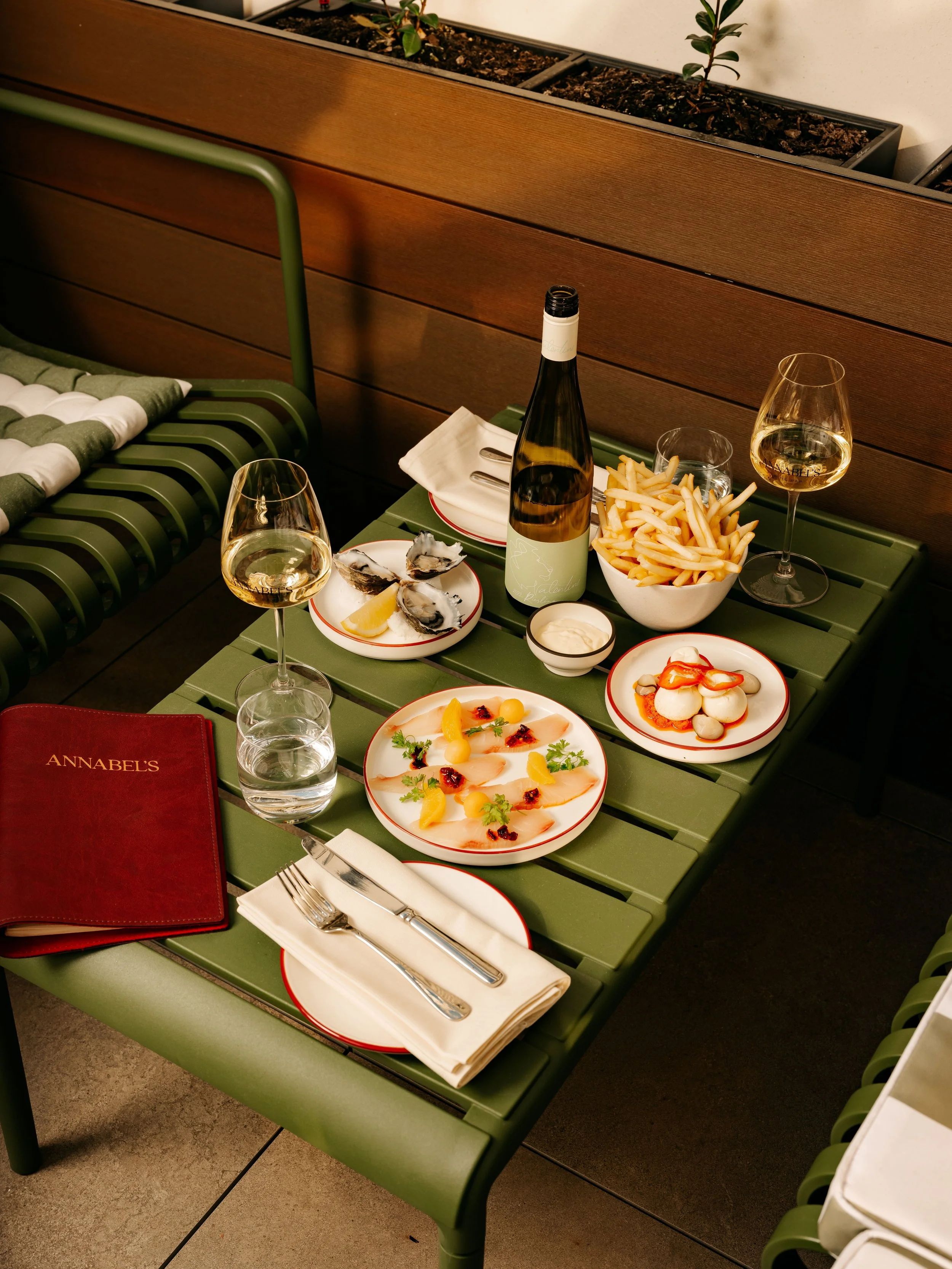 A table set with plates of oysters, sliced vegetables, shoestring fries, two glasses of white wine, a bottle of wine in Annabel's Patio