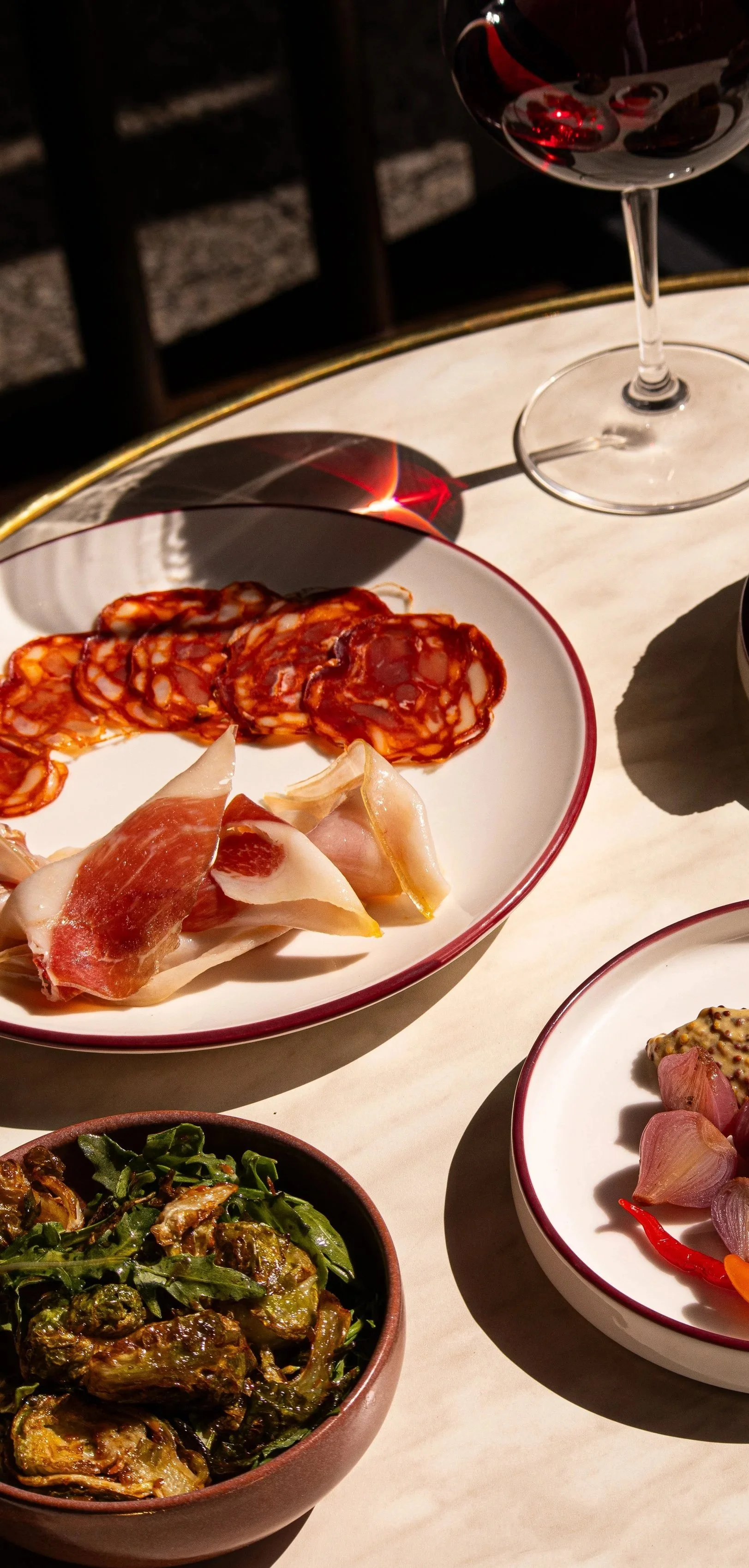 A white plate with assorted cured meats, including slices of salami and prosciutto, placed on a table next to a glass of red wine at Annabel's Wine Bar