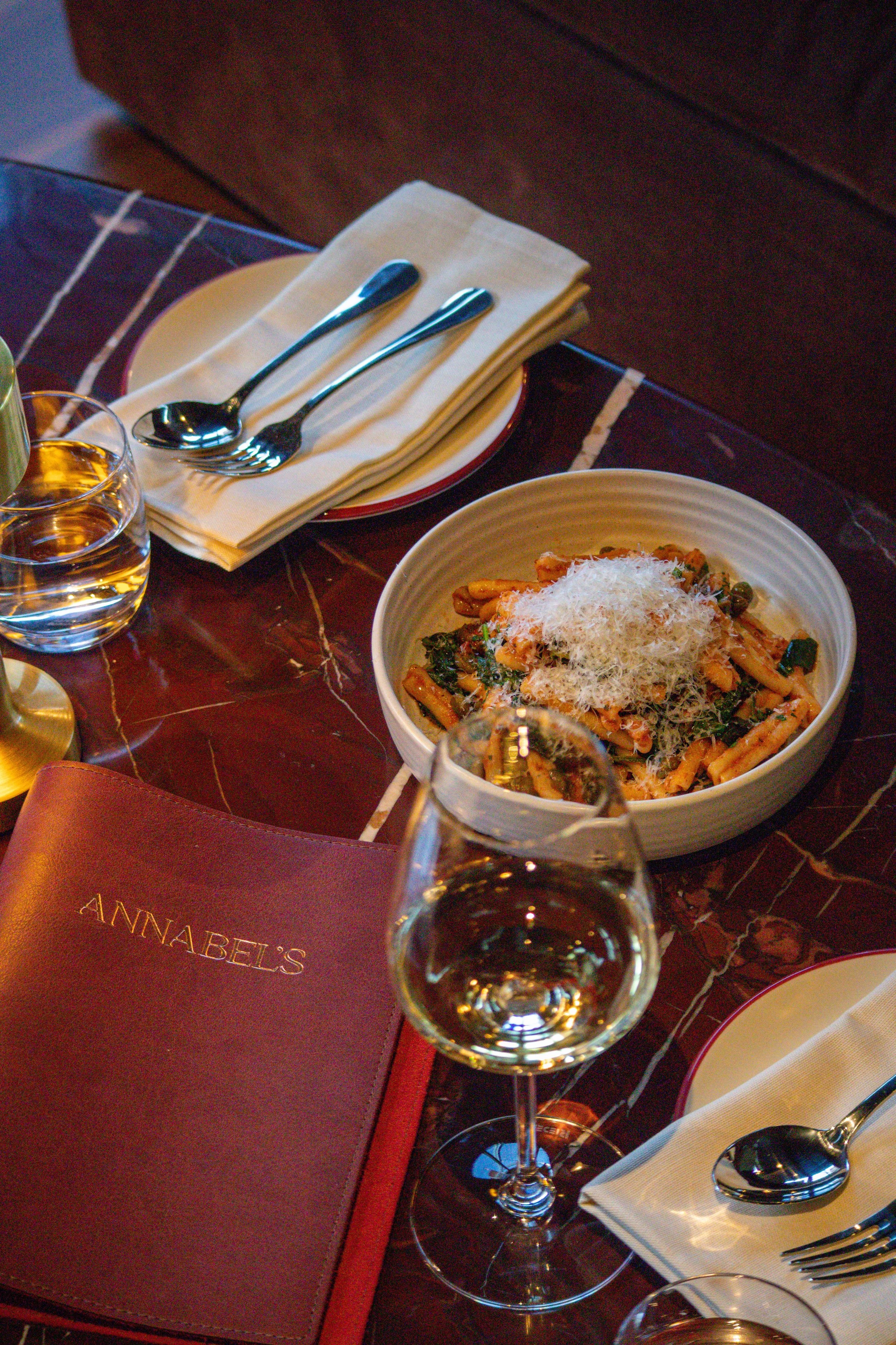 A table with a bowl of pasta topped with grated cheese, a glass of white wine, silverware, a rolled napkin, and a menu labelled 'Annabel's'. On the Pasta and Vino Night on Wednesday