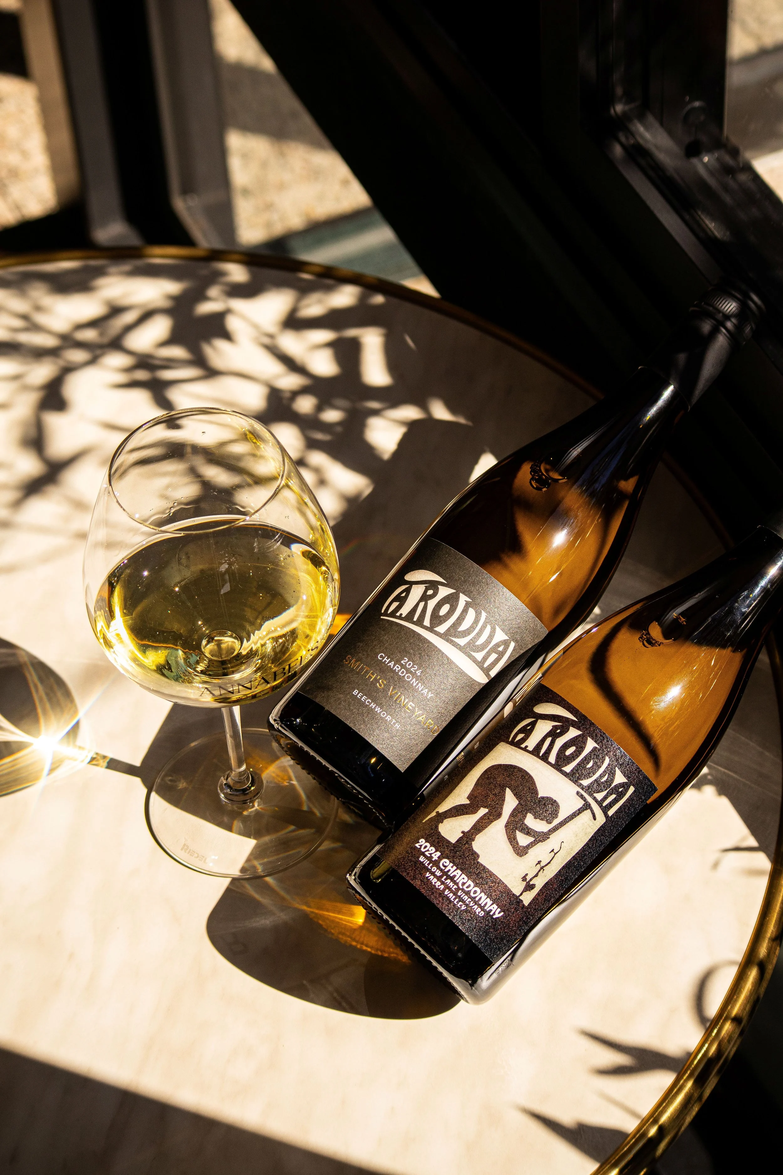A glass of white wine and two bottles of Chardonnay wine on a white table, casting shadows with leaf patterns, with sunlight highlighting their presence.