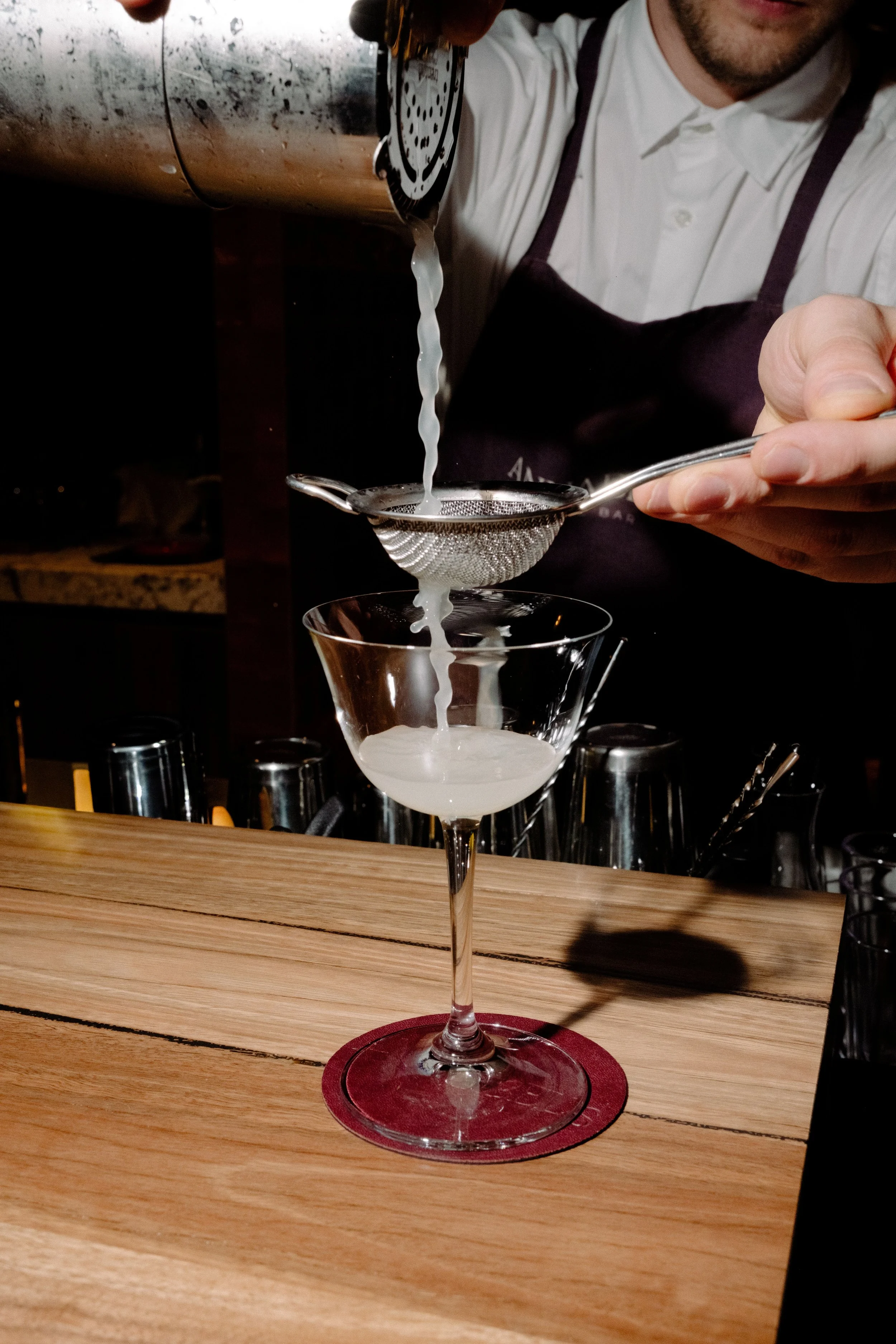 An Annabel's bartender pours a martini into a cocktail glass, with a wooden bar counter and barware in the background