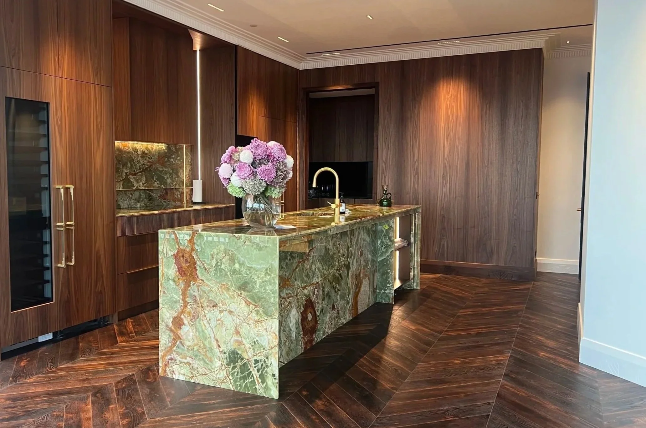 Modern kitchen with a green marble island, a flower vase with purple and white flowers, wooden cabinets, and a herringbone dark wood floor.