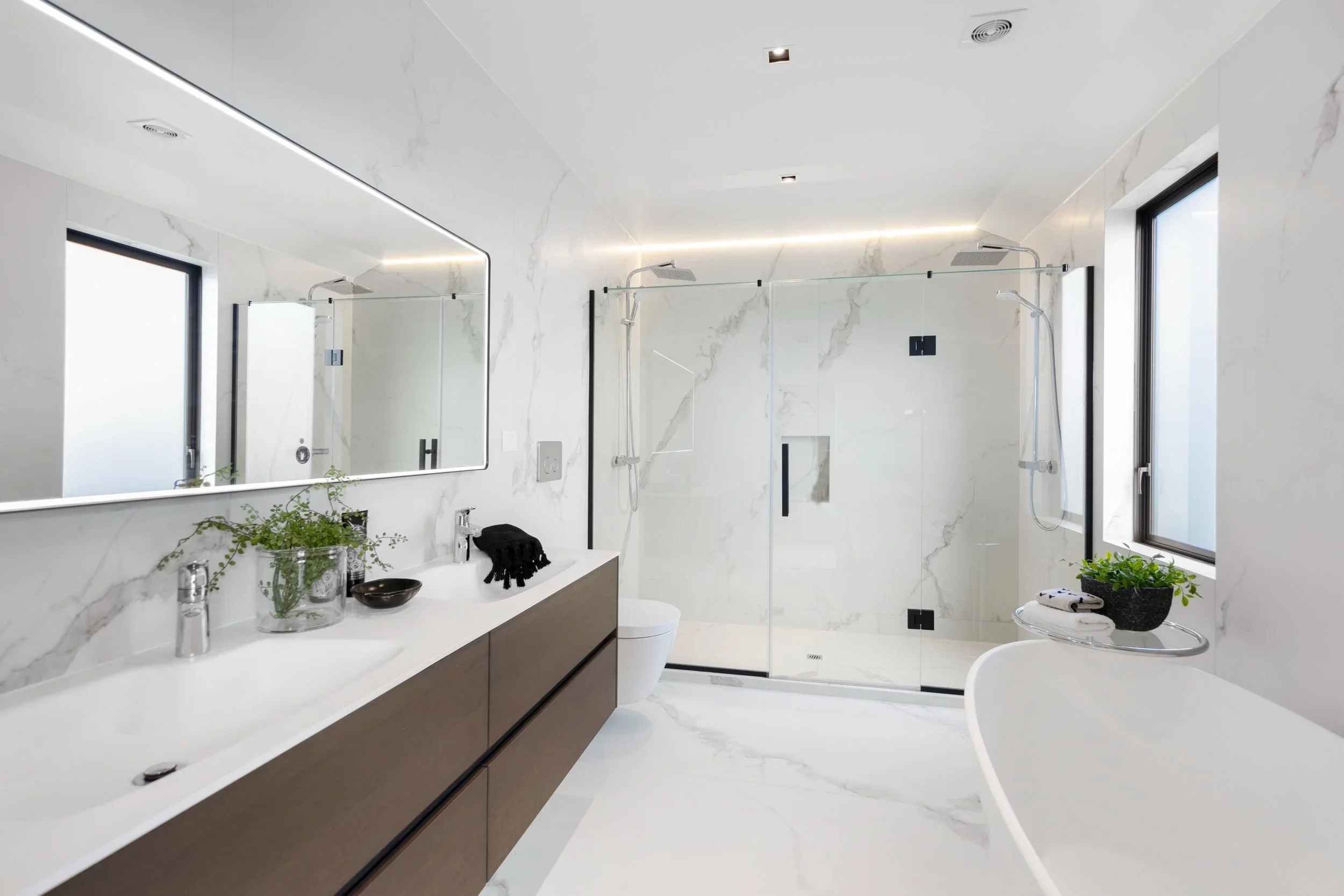 Modern bathroom with white marble walls and floors, a large mirror, double sink vanity, a glass shower enclosure, a freestanding bathtub, and black-framed windows with plants on the counter and bathtub