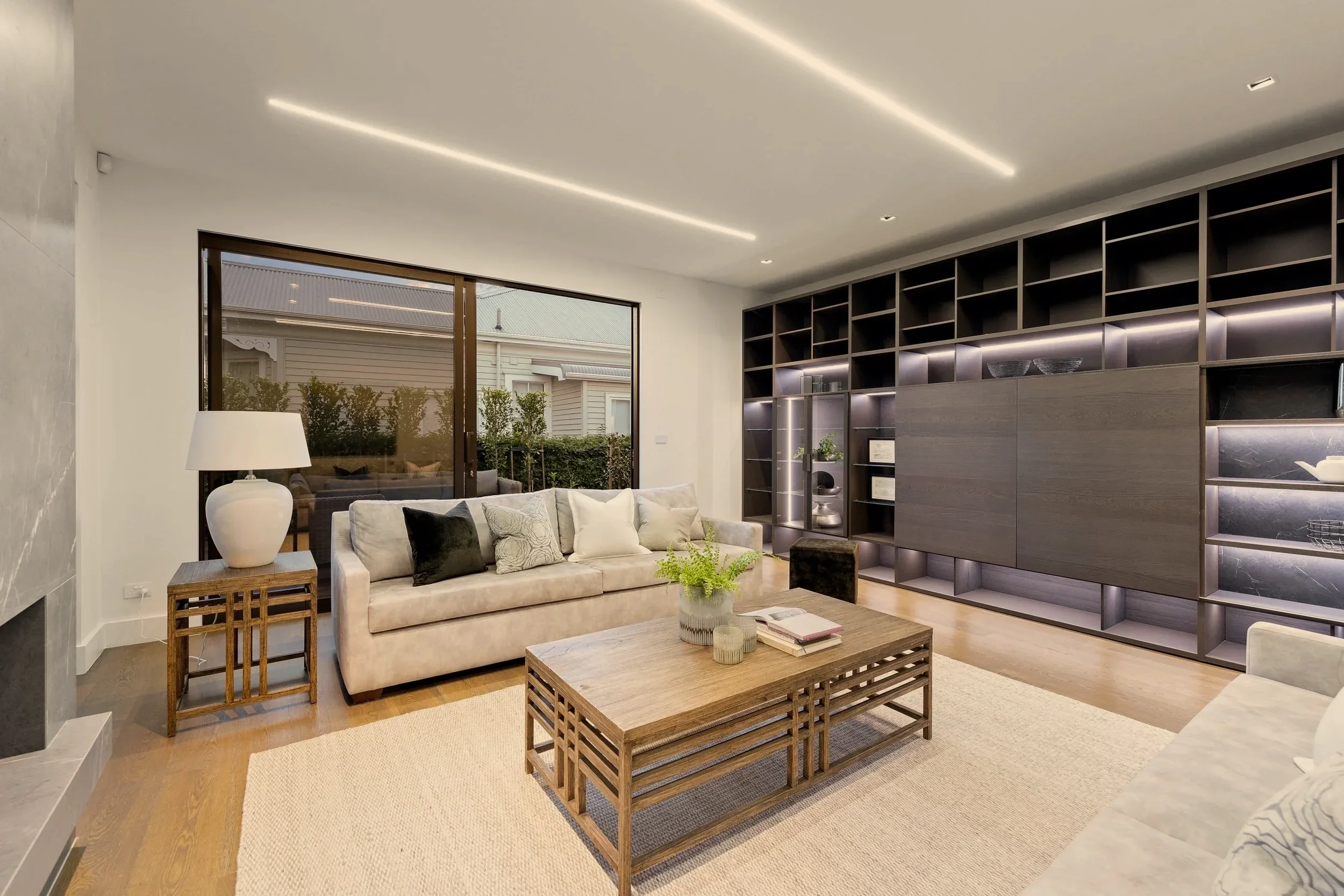 Modern living room with beige sofa, wooden coffee table, and built-in dark shelving unit. A large window shows a view of the outside garden, and a lamp is on a small wooden side table.