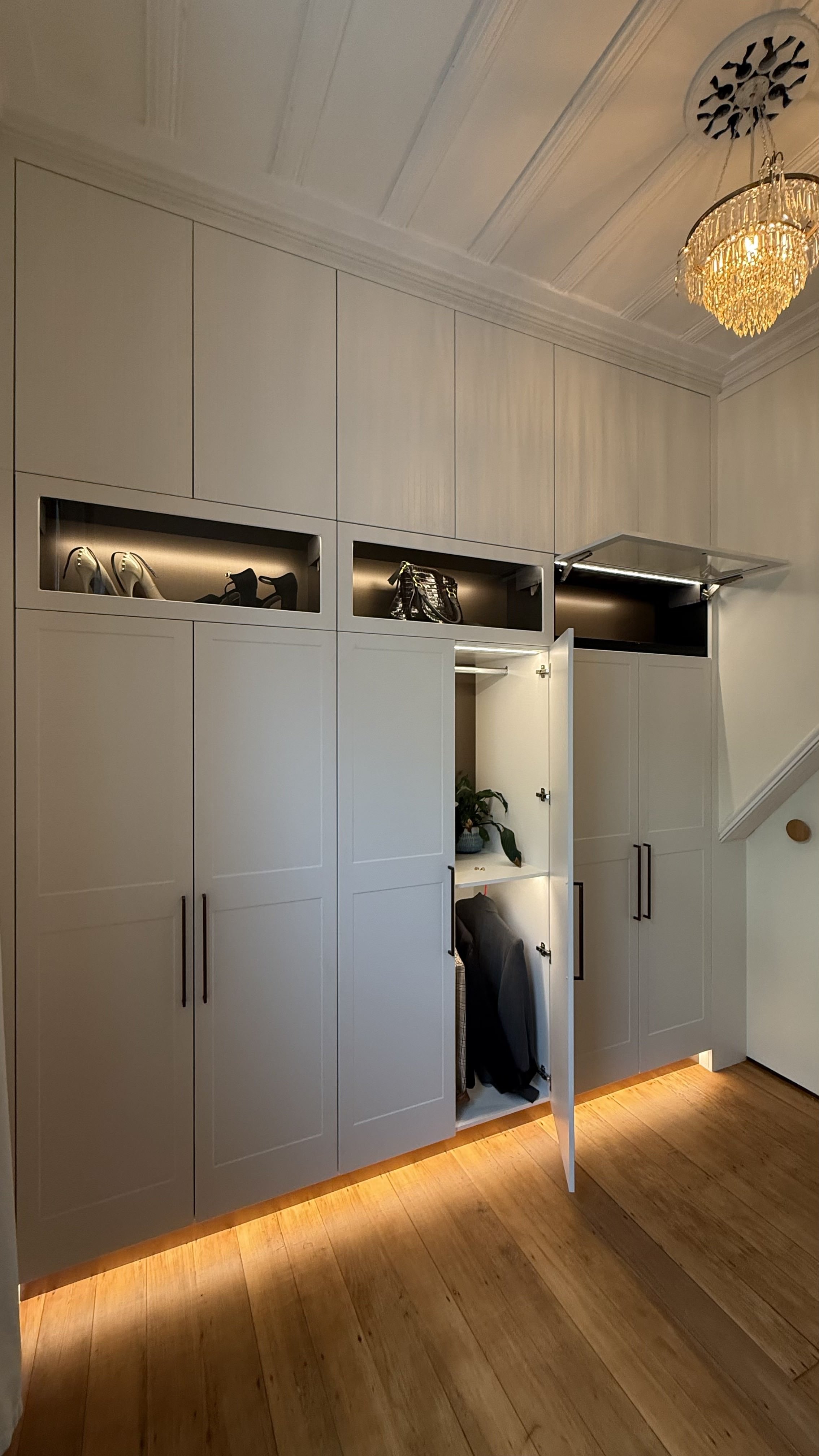 Built-in white storage cabinets with open compartments holding high-heeled shoes, handbags, and a coat inside open cabinet doors. Wooden flooring and a chandelier hanging from the ceiling.