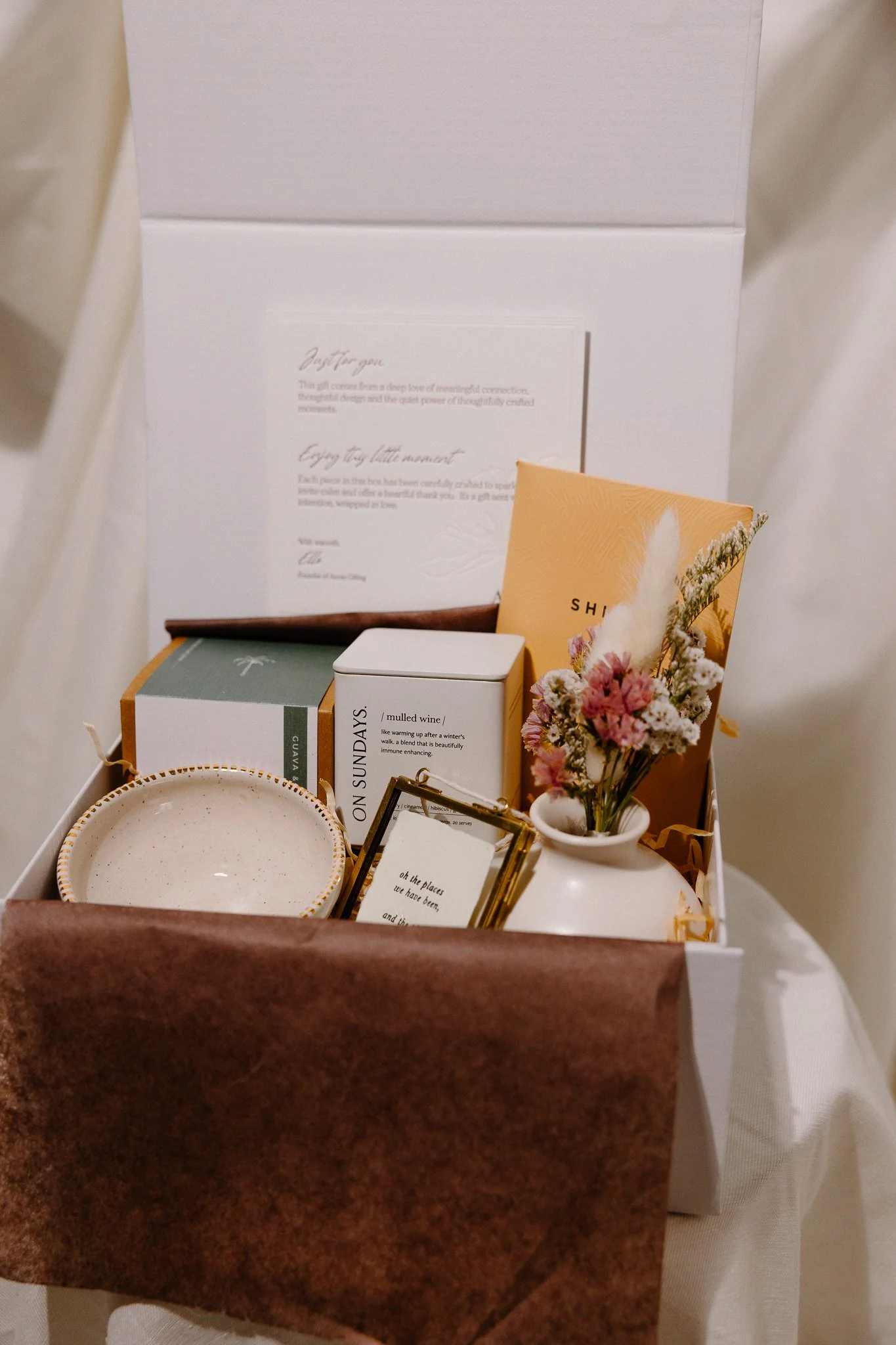 A white gift box with a dark green satin ribbon tied in a bow, sitting on a white surface with a plant in the background.