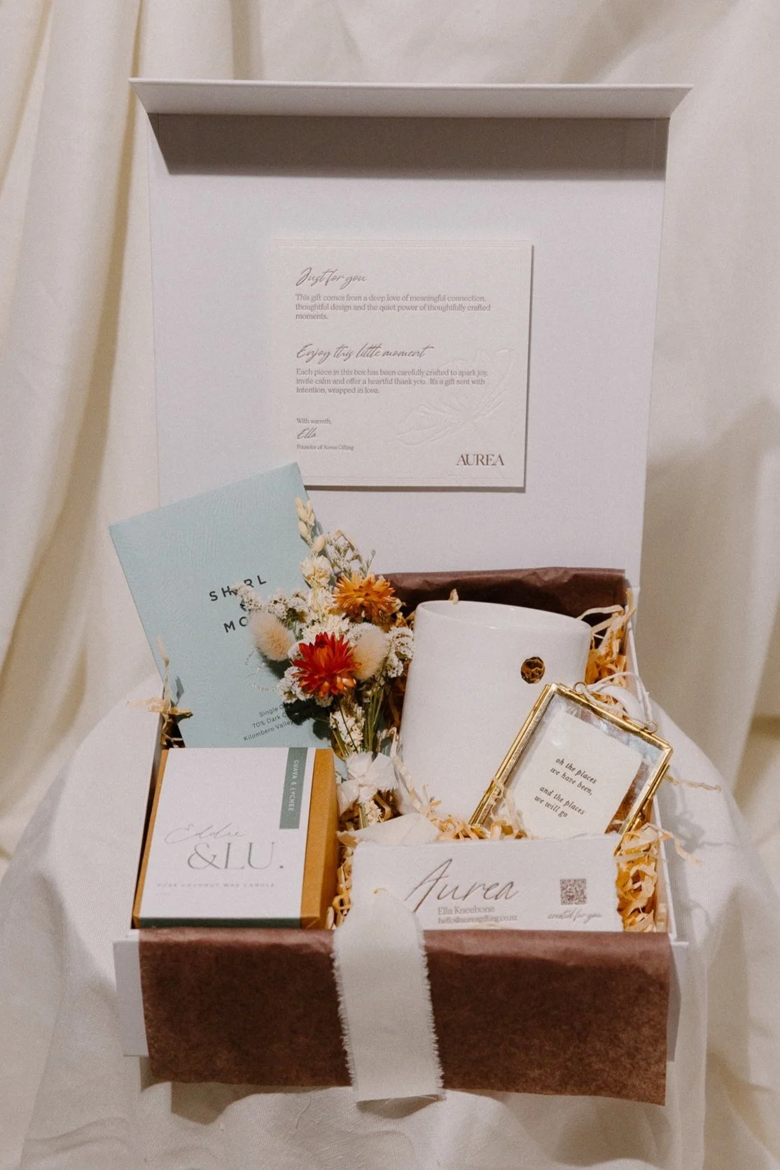 A white gift box wrapped with a dark red ribbon and bow, placed on a table next to an orange greeting card and matching envelope, with a dark leafy plant in the background.