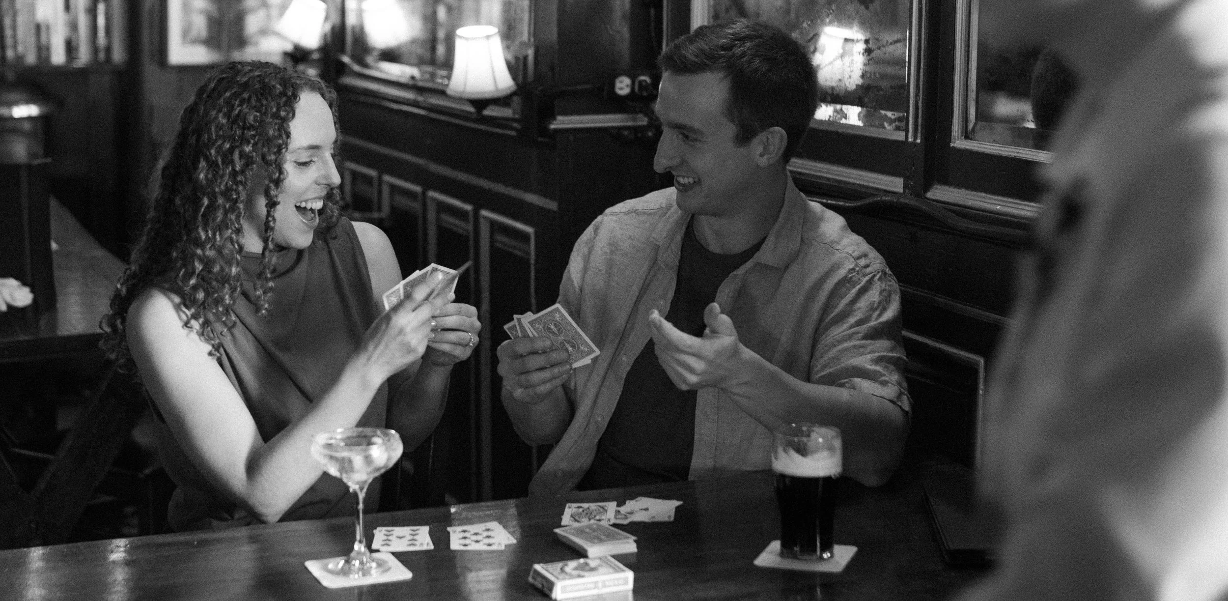 Two people, a woman with curly hair and a man, sitting at a table in a bar or pub, playing cards and laughing together. The woman is holding cards, and the man is holding cards with a glass of dark beer and a cocktail nearby.