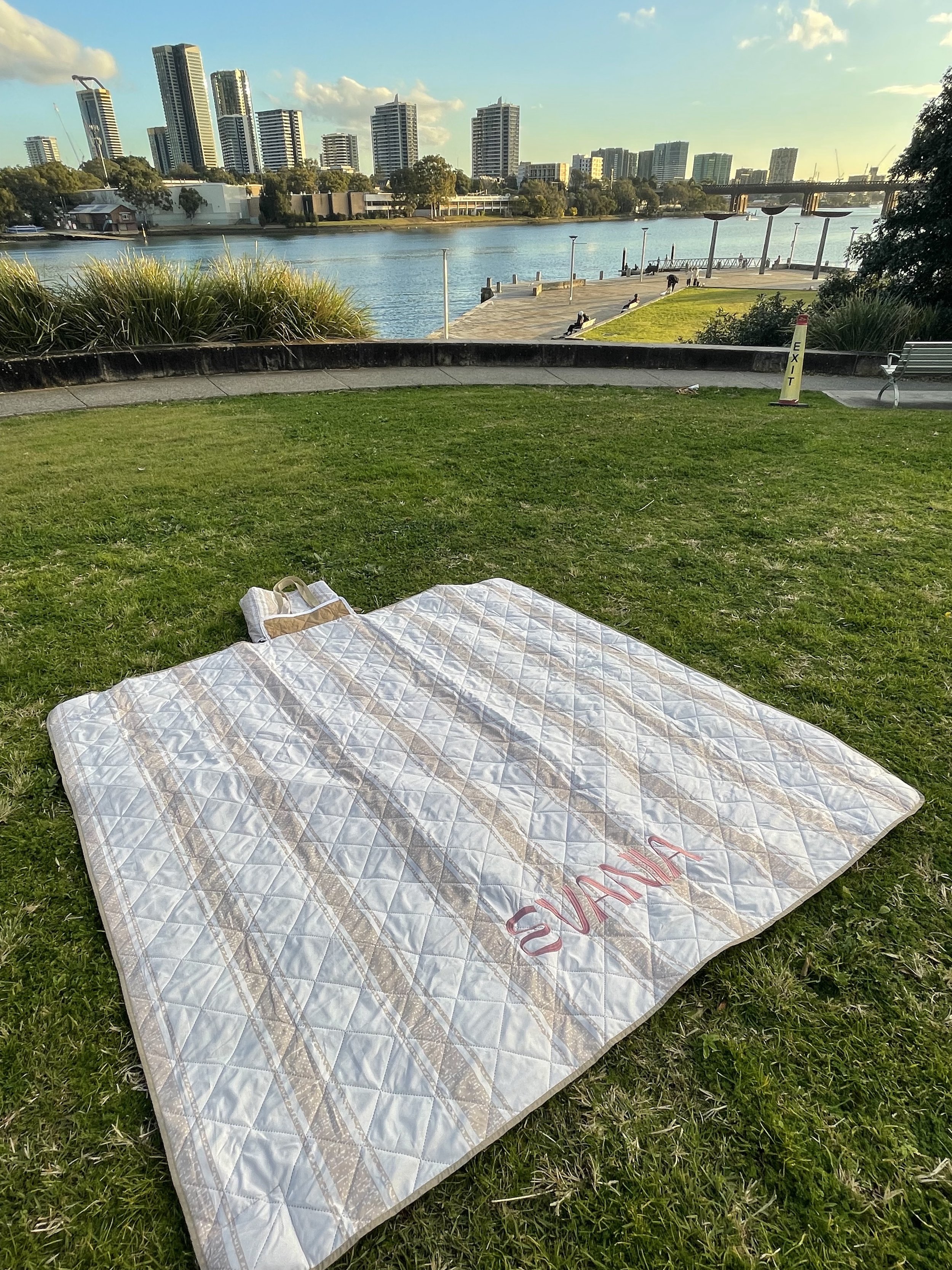 A blanket with the word "SUNSET" embroidered on it is laid out on the grass in a park near a river with a city skyline in the background. There are some benches and a yellow buoy marked "EXIT" nearby.