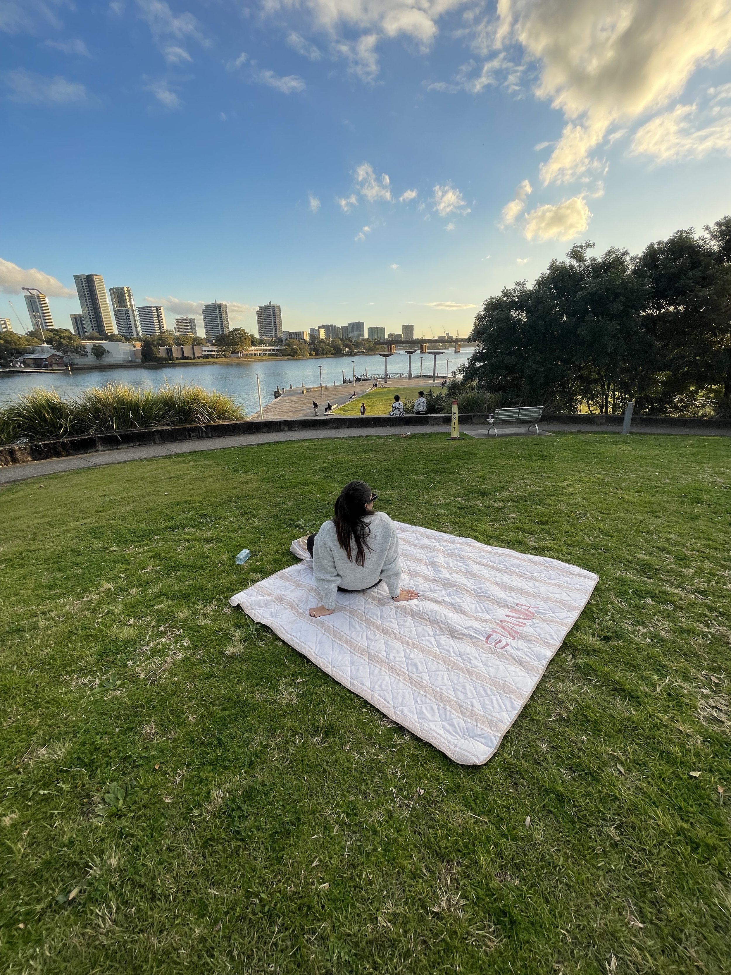 A woman sitting on a white blanket on grass in a park by a river, with city buildings in the background on a sunny day.
