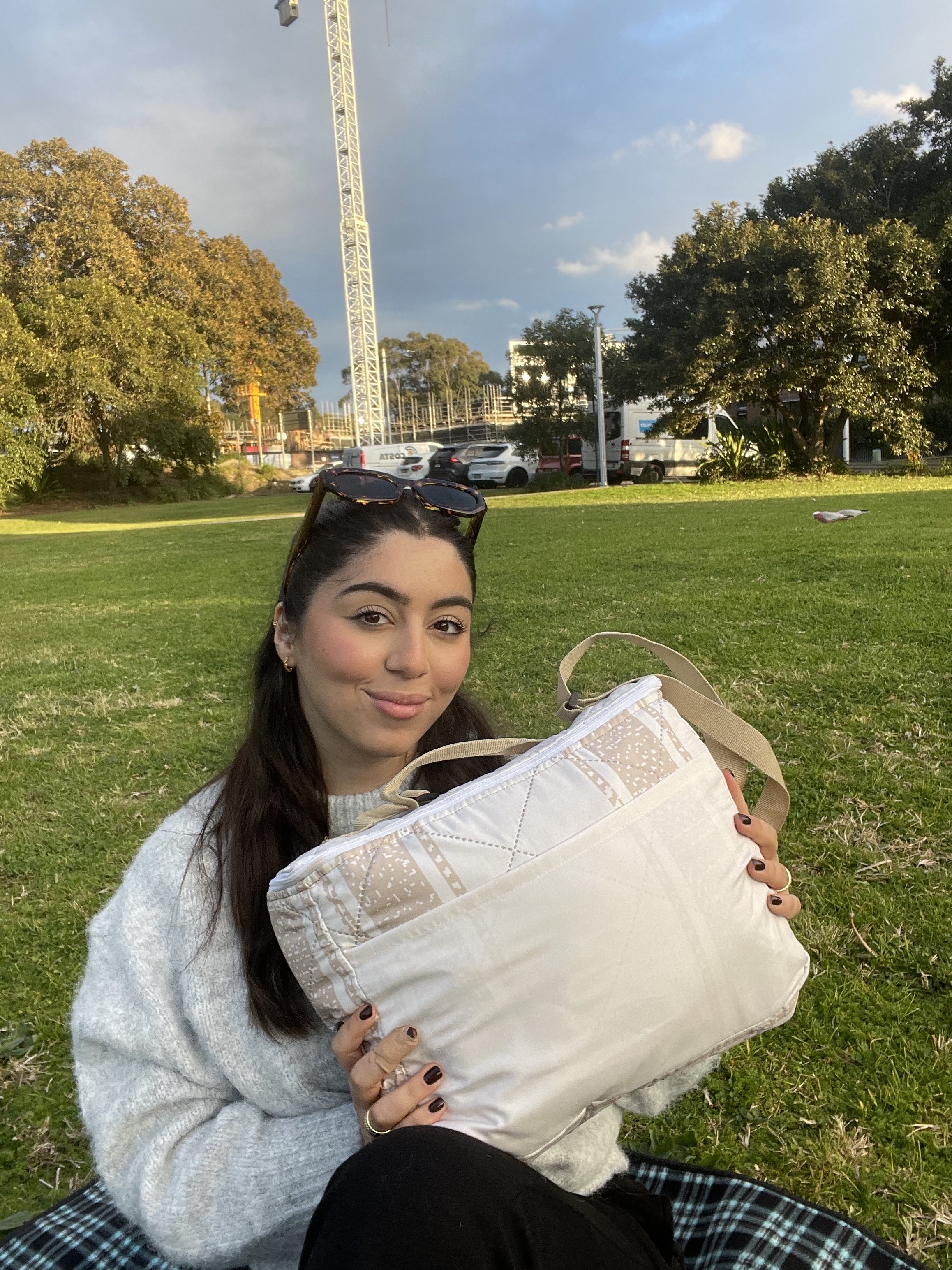 Young woman sitting on grass in park, holding a beige bag, with sunglasses on her head, smiling at camera.