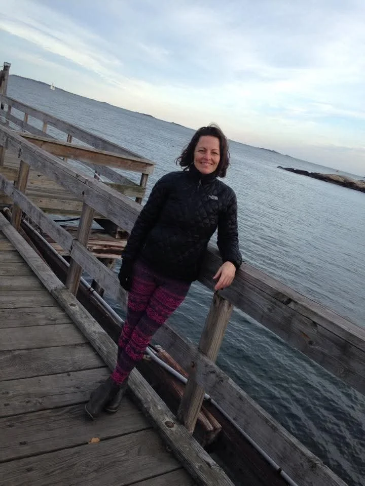 Woman standing on a wooden pier by the water, smiling, with a shoreline and cloudy sky in the background.