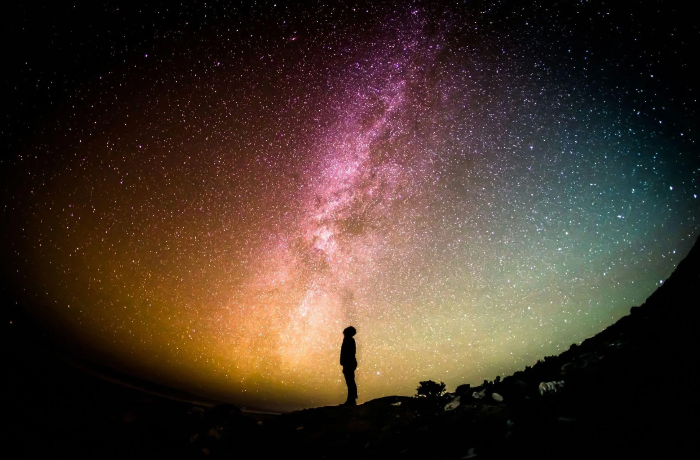 A person standing on a hill at night, gazing at a colorful, star-filled sky with the Milky Way galaxy visible.
