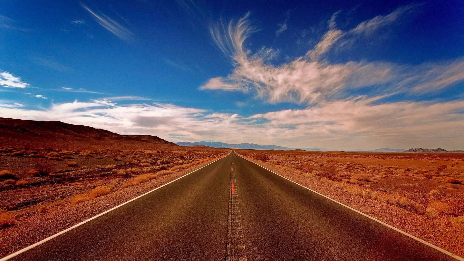 A straight rural highway stretching into the horizon through a desert landscape with mountains in the distance, under a partly cloudy sky.