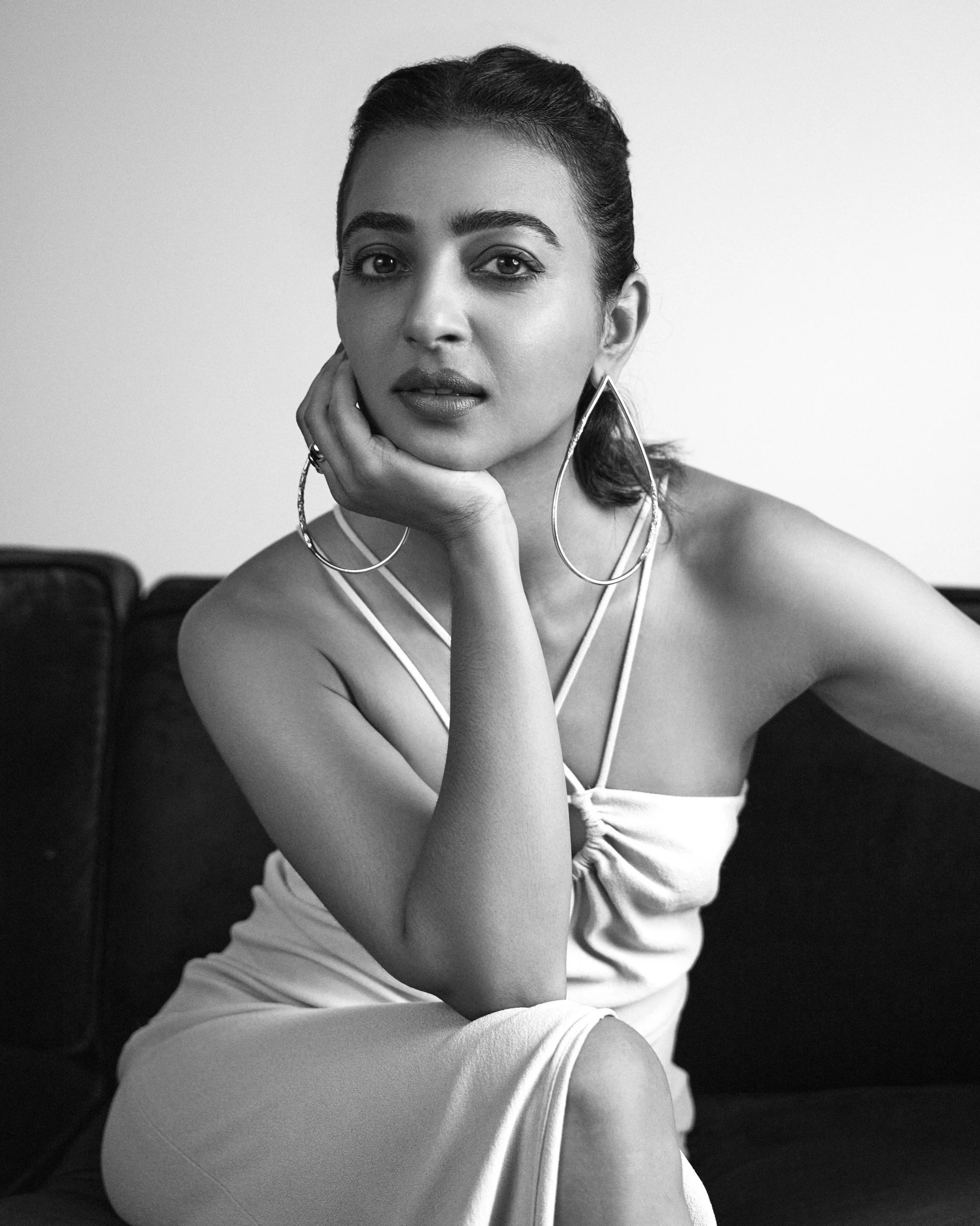 Black and white portrait of a woman with dark hair, wearing large hoop earrings and a light-colored dress with thin straps, sitting on a sofa with a thoughtful expression.