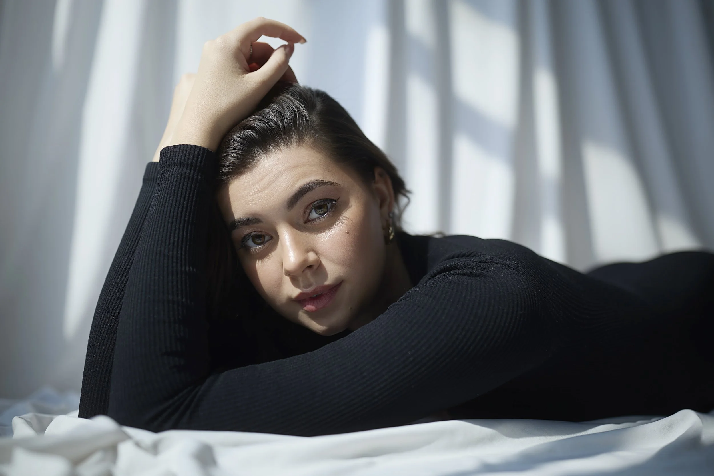 A young woman with brown hair, lying on her stomach on a bed, resting her head on her arm, with sunlight filtering through white curtains in the background.