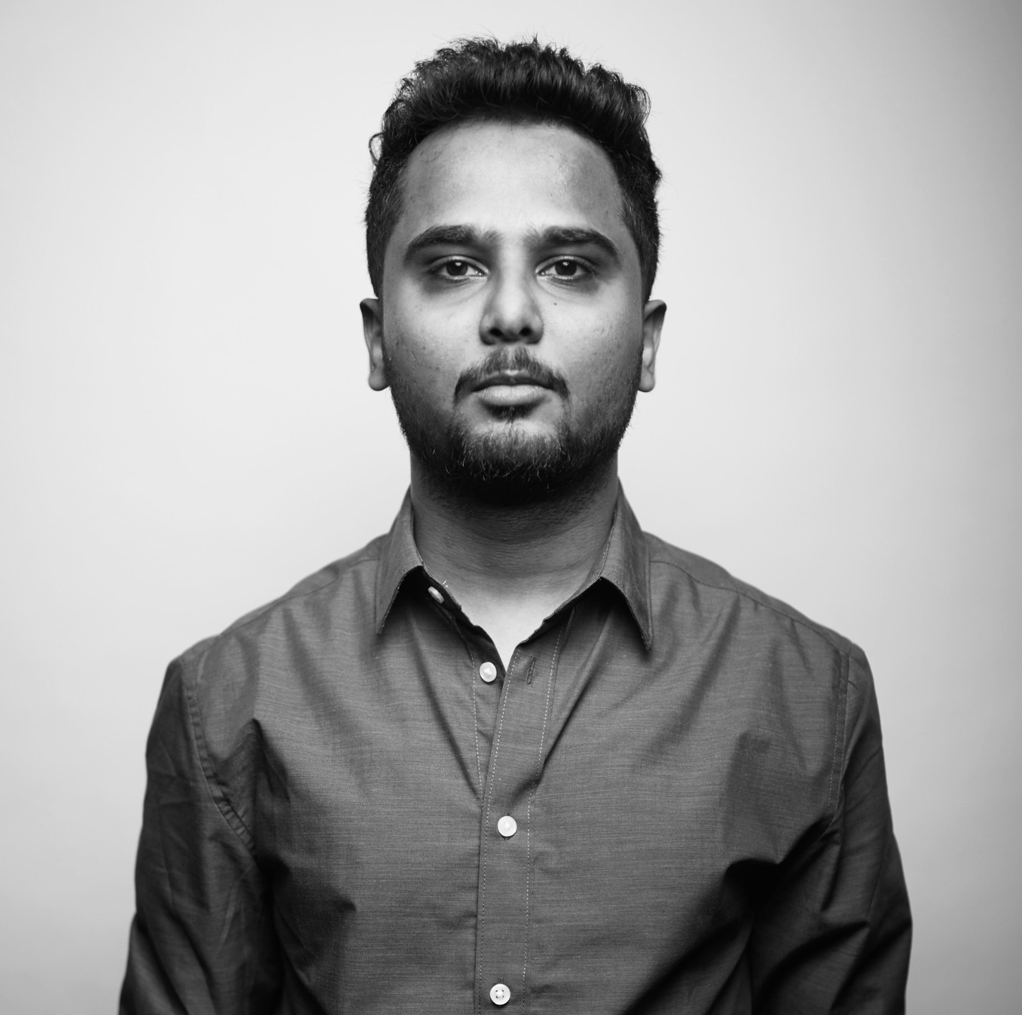 Black and white portrait of a young man with dark hair, beard, and mustache, wearing a button-up shirt, looking directly at the camera against a plain background.