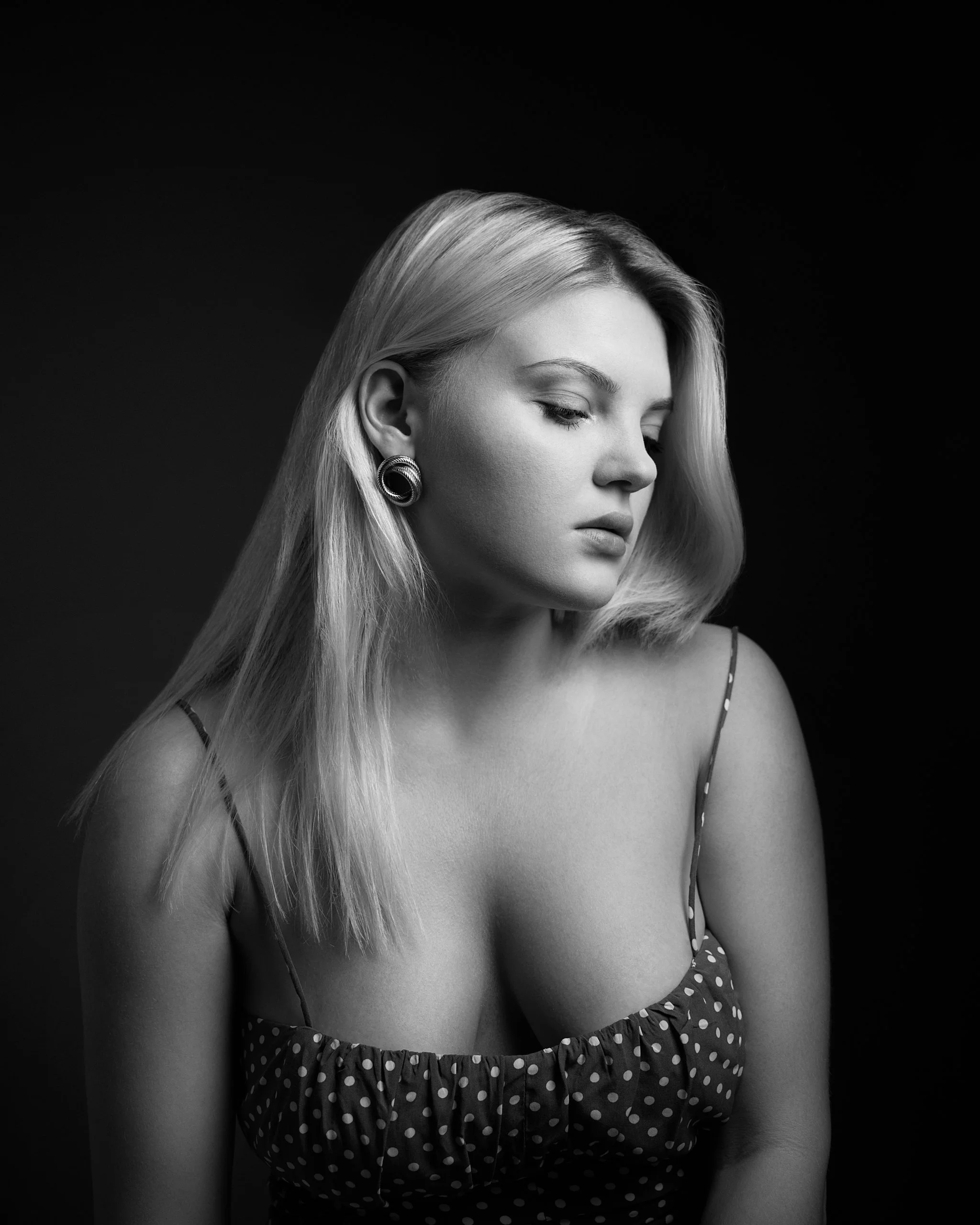 Black and white portrait of a young woman with long blonde hair, wearing earrings and a polka-dot dress, looking down with a contemplative expression.