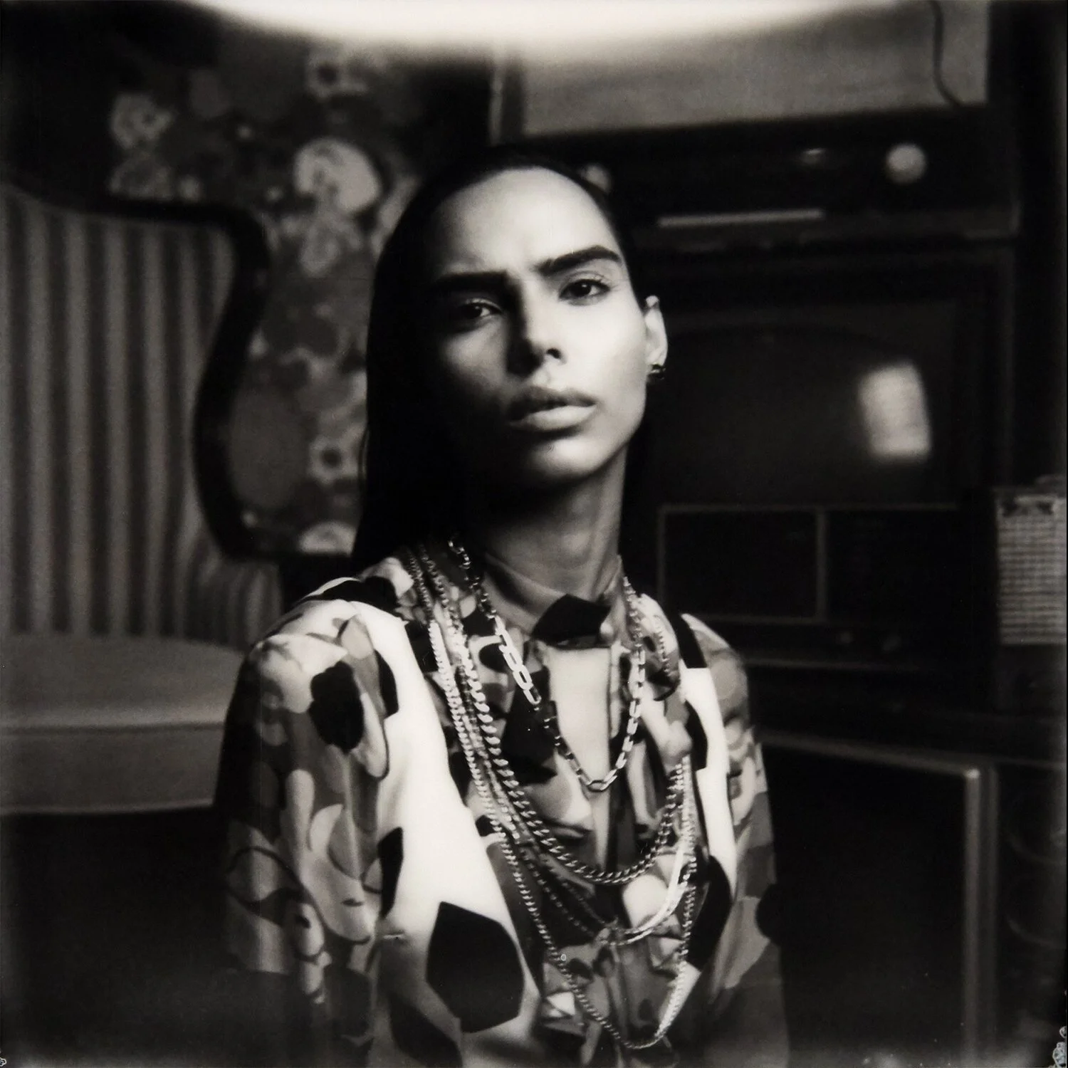 Black and white photo of a woman wearing multiple chains and a patterned shirt, with a serious expression, in a room with a vintage television and other old-fashioned furniture.