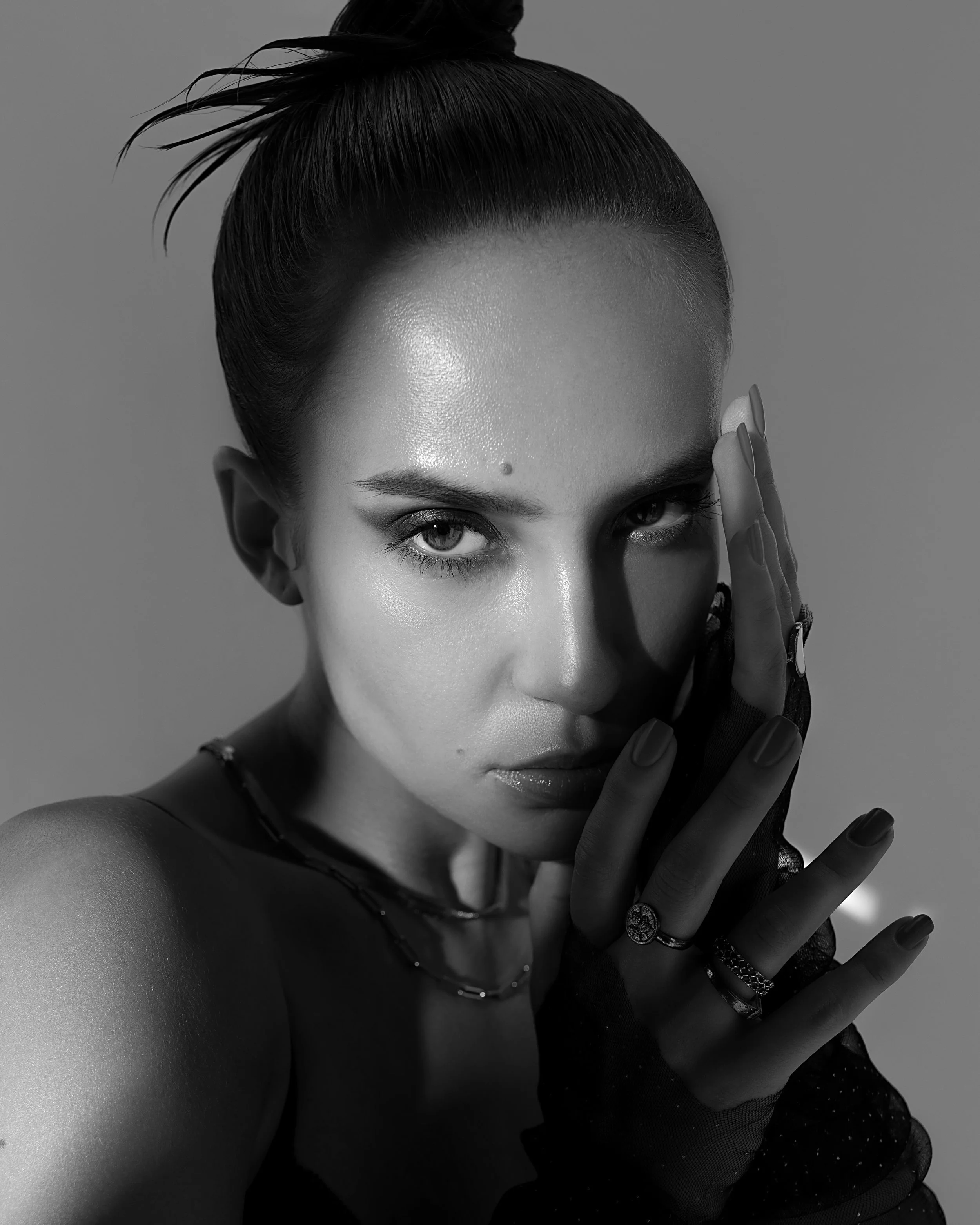 Black and white portrait of a woman with intense eyes, pulled-back hair, and wearing rings and rings on her fingers, touching her face in a dramatic pose.