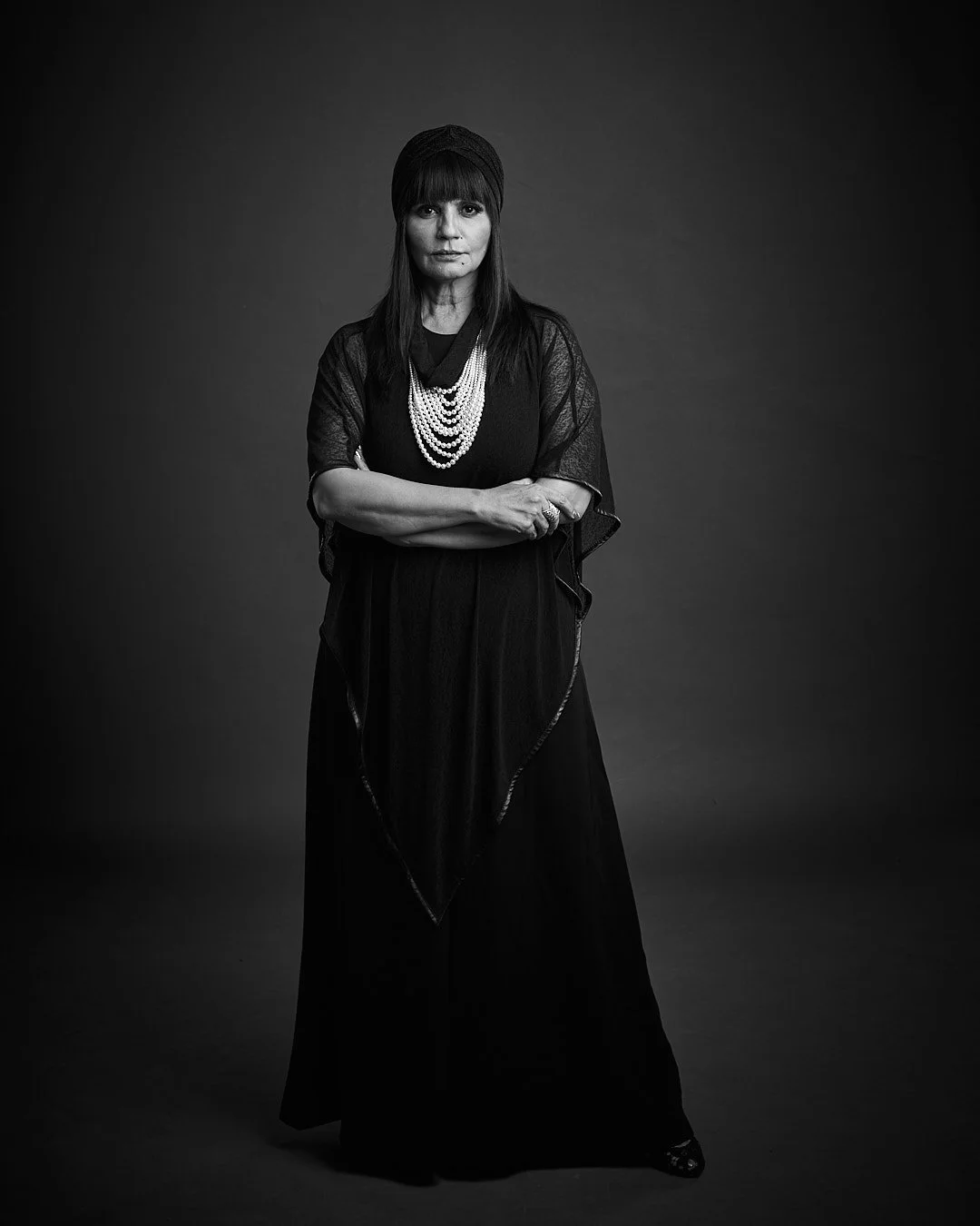 A black and white photo of a woman wearing a head wrap, layered pearl necklaces, and a long black dress, standing with her arms crossed against a dark background.