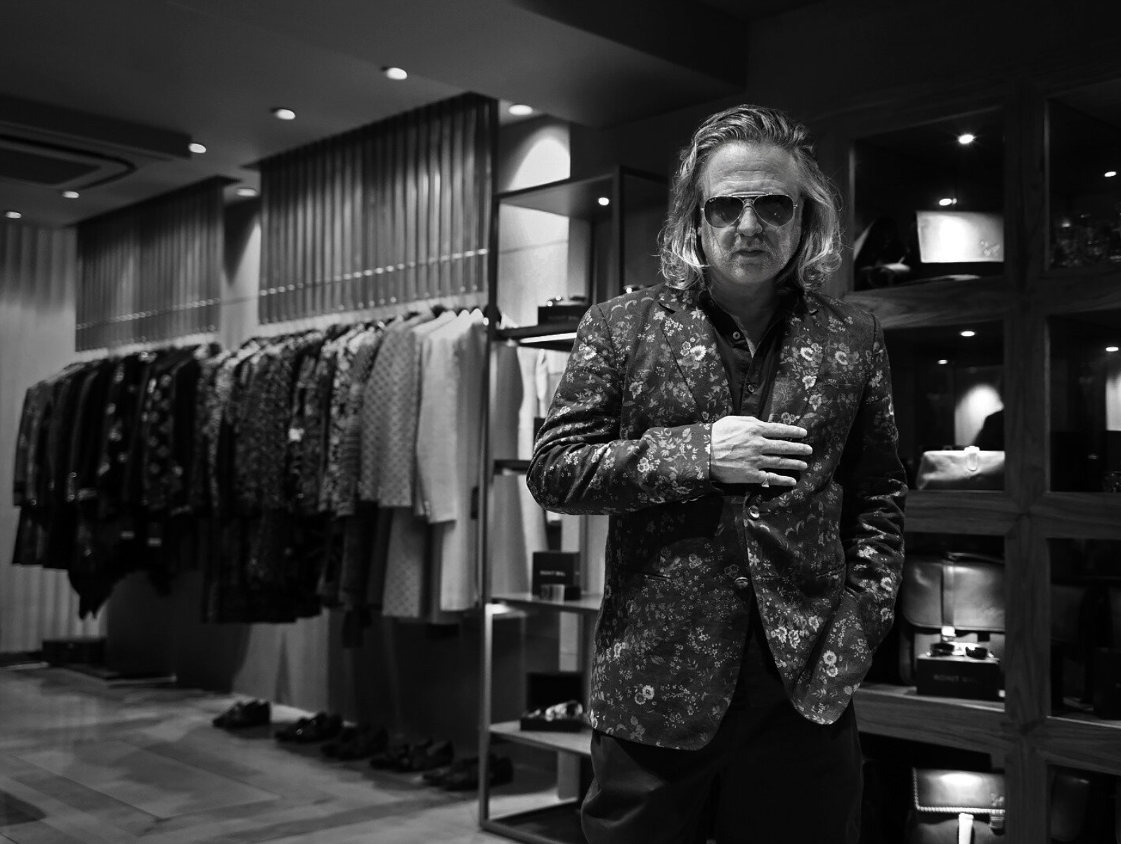 Black and white photo of a man with long hair, wearing sunglasses and a floral-patterned blazer, in a clothing store with racks of shirts and pants in the background.