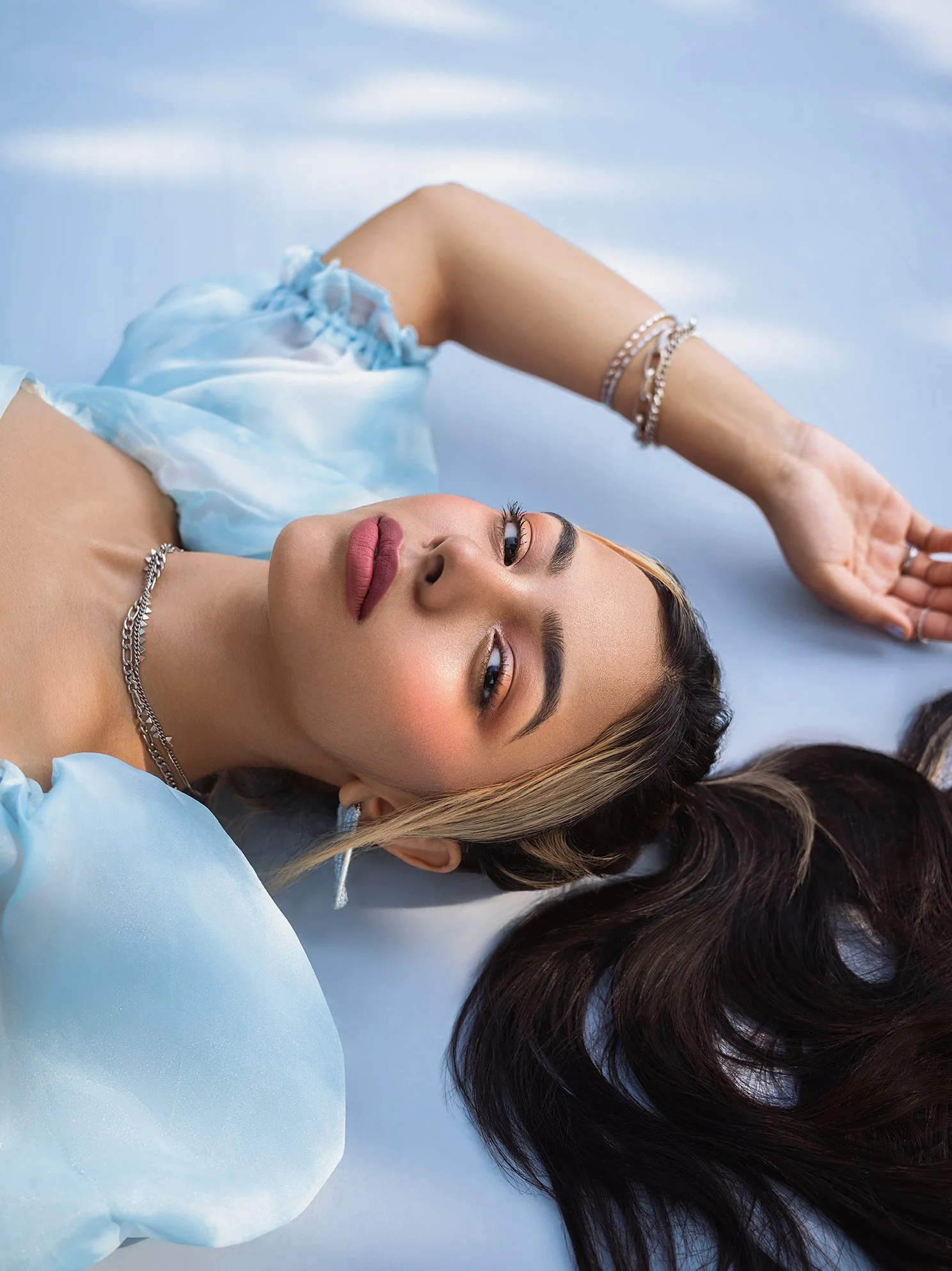 A woman lying on a light blue surface, with her head turned slightly to the side, wearing a light blue satin blouse and jewelry, with dark brown hair spread out.