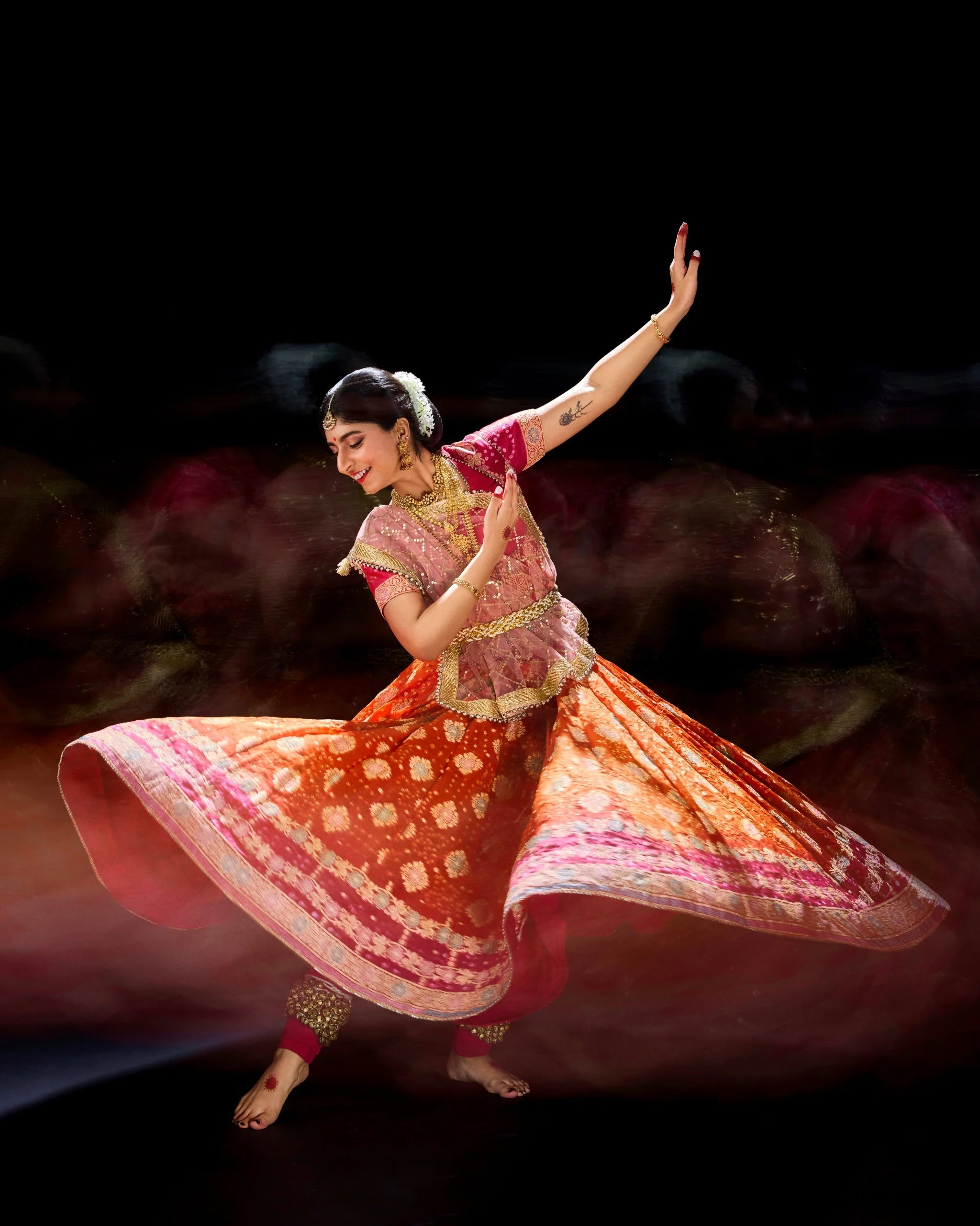 A woman in traditional Indian attire performing a classical dance, wearing a pink and orange embroidered outfit, jewelry, and ornaments, with a black background.