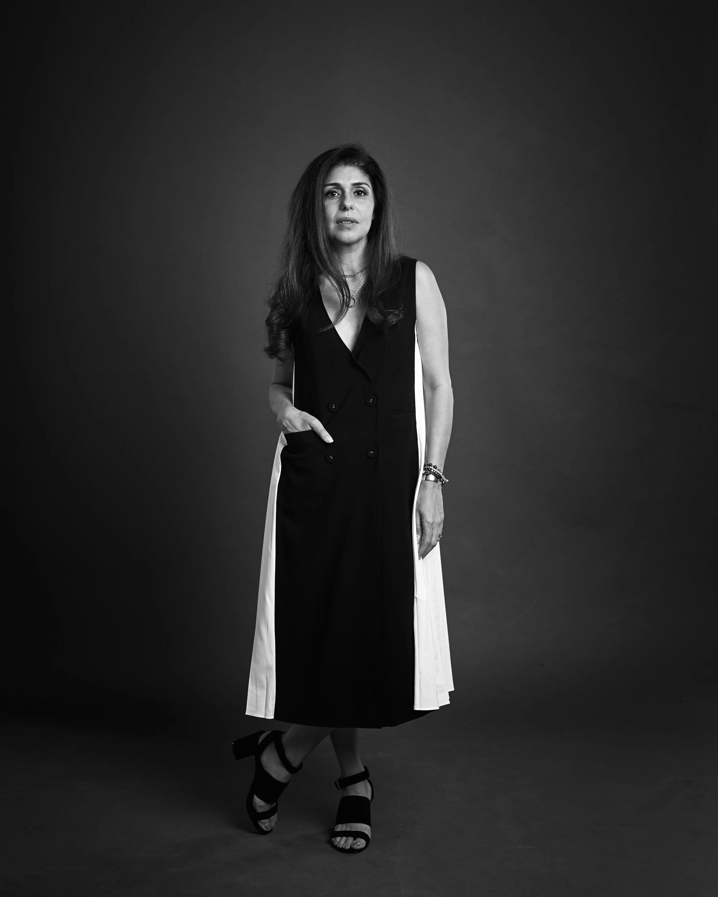 A woman with long hair wearing a sleeveless black and white dress, standing with one hand in her pocket, against a plain dark background.