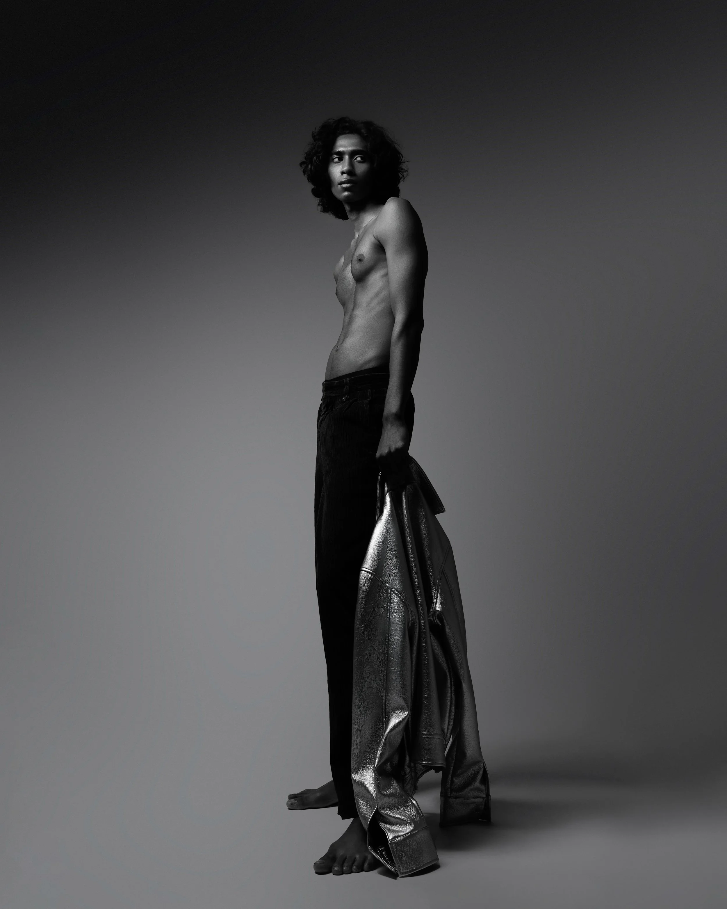A black and white photo of a topless person with curly hair holding a leather jacket, standing barefoot against a gradient background.