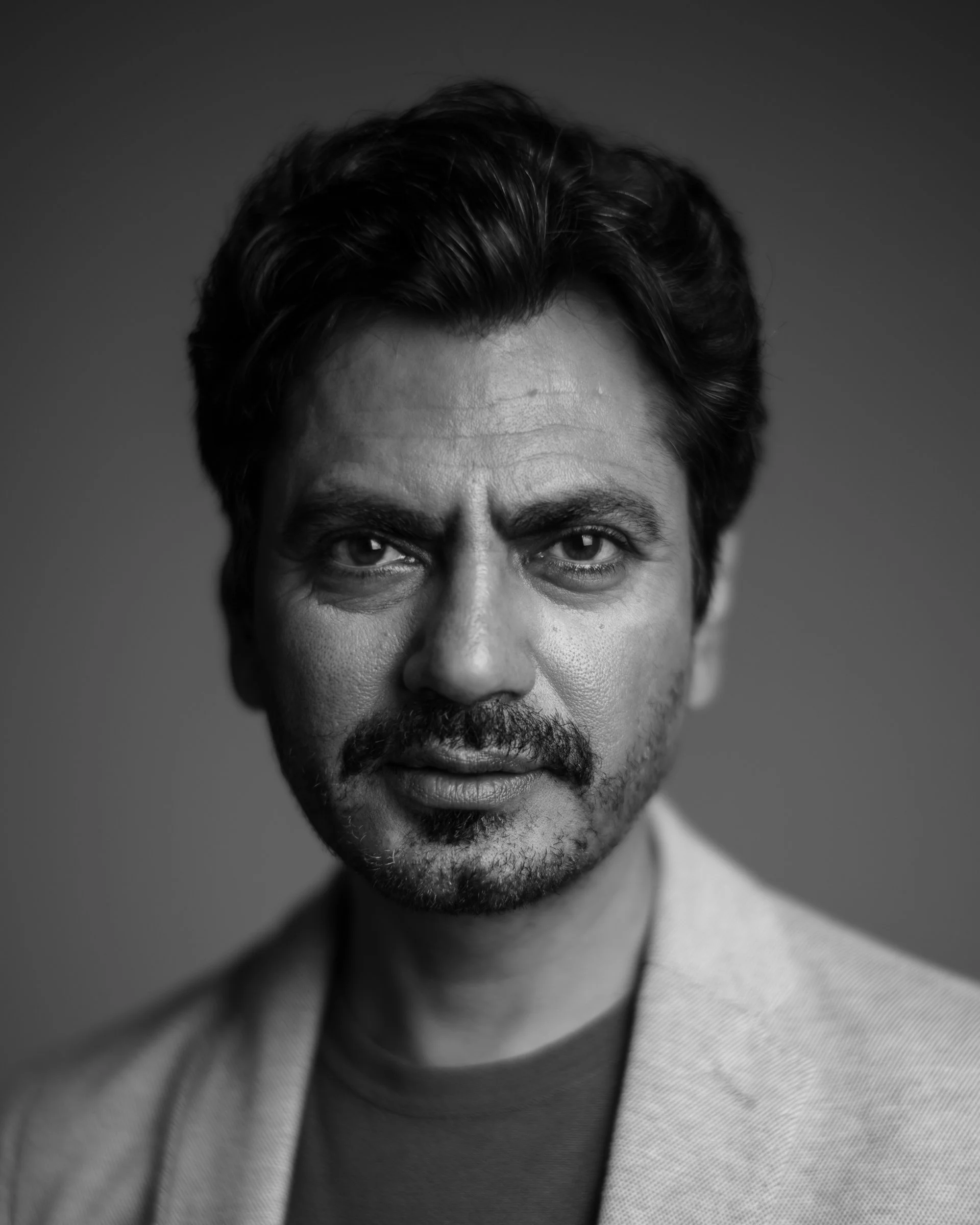 Black and white close-up portrait of a man with dark hair, beard, and mustache, wearing a light-colored blazer and a dark shirt, looking directly at the camera with a serious expression.