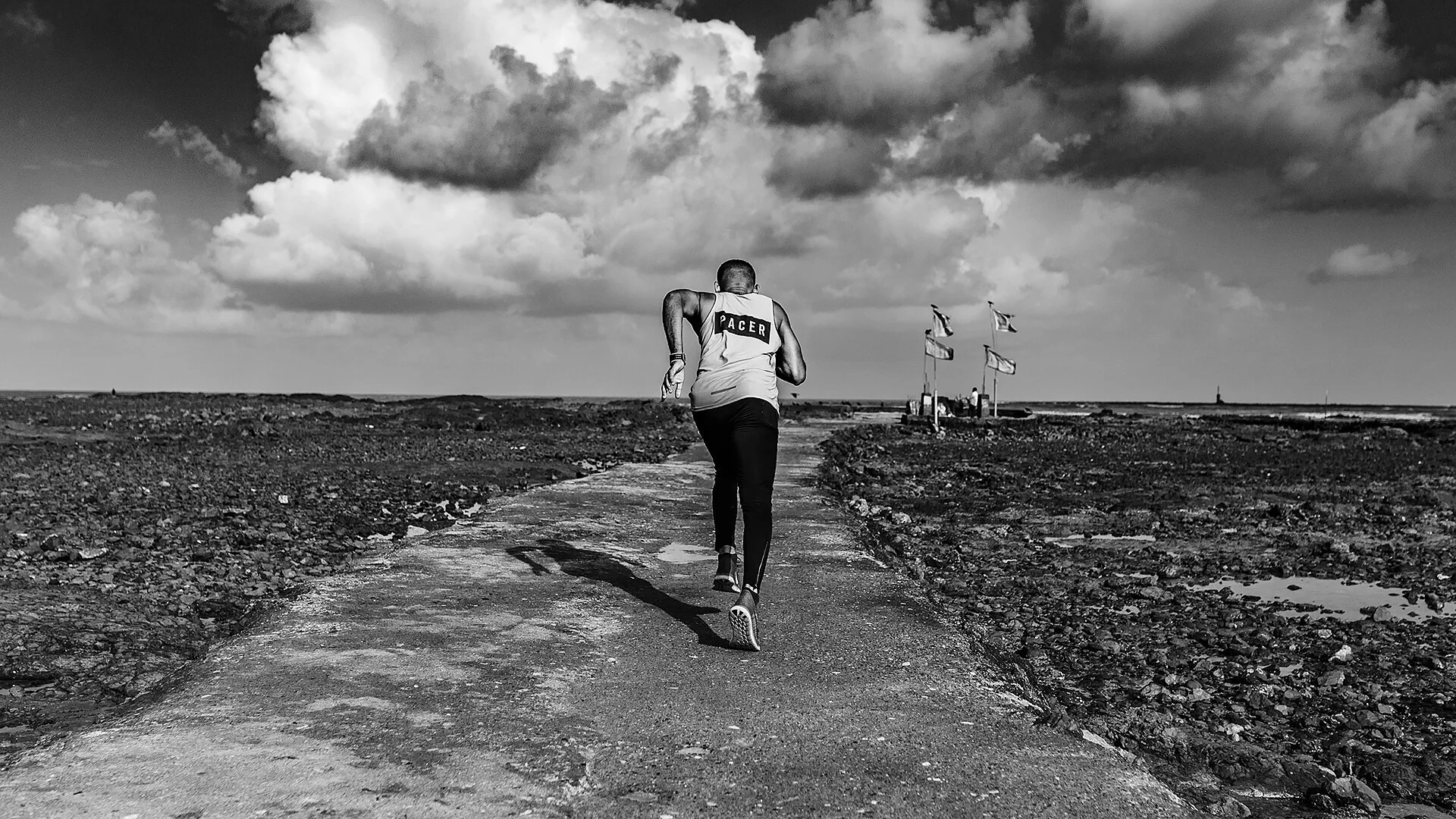 A man running on a paved path in a vast, open landscape with dark clouds overhead. The man is dressed in athletic clothing, wearing a tank top with the word "PACER" on the back.