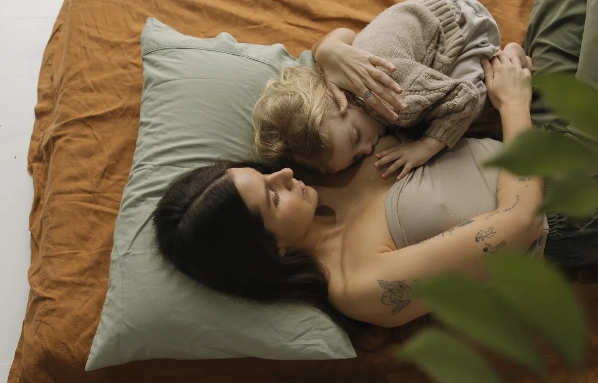 A woman with black hair lying on a bed with a light green pillow, with a young girl with blonde hair lying on her chest, resting peacefully. The woman has tattoos on her arm, and the girl is wearing a brown sweater. Breastfeeding a toddler.