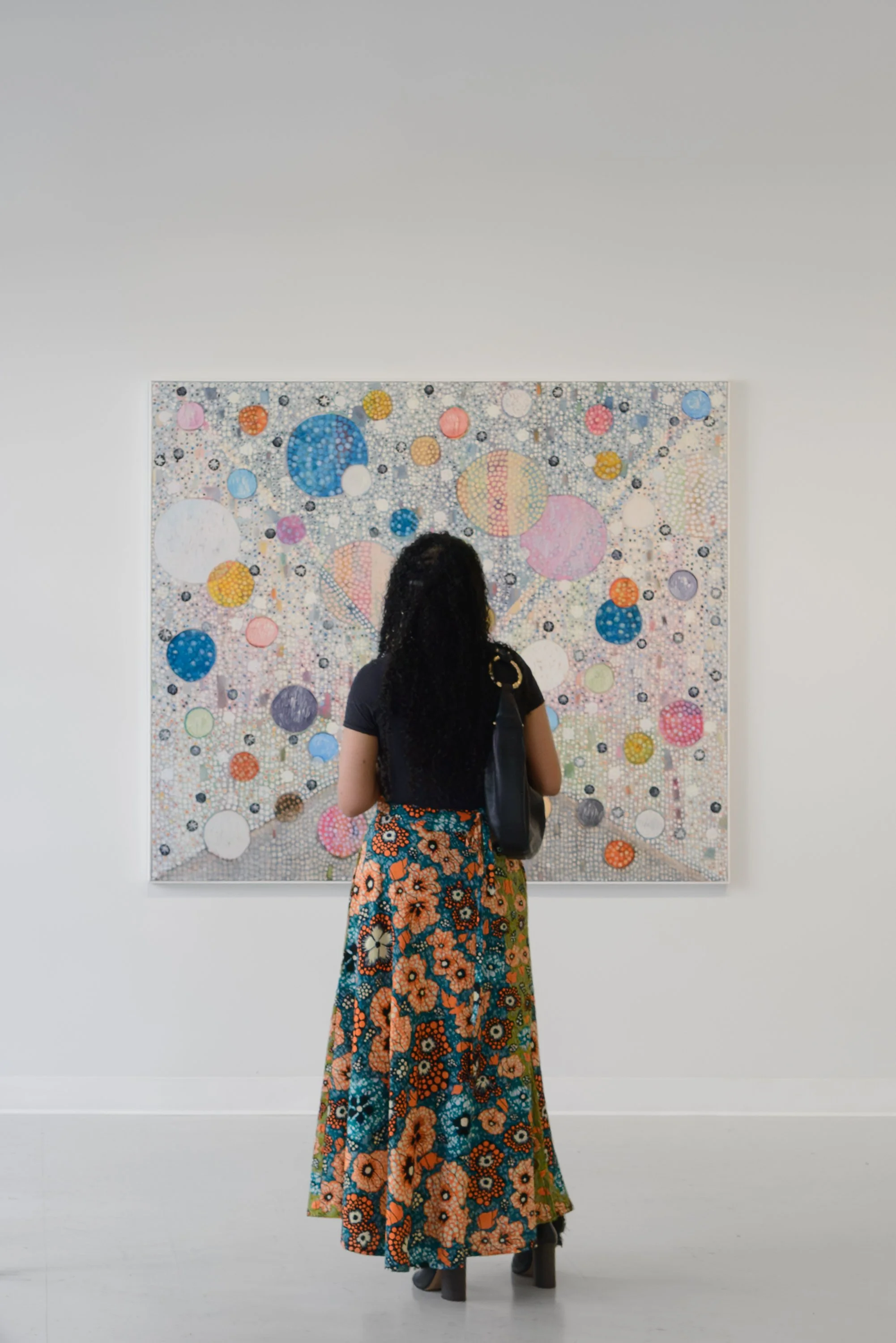 A woman with long black curly hair, wearing a black top and a colorful floral maxi skirt, stands facing an abstract bubble art piece at an art gallery.
