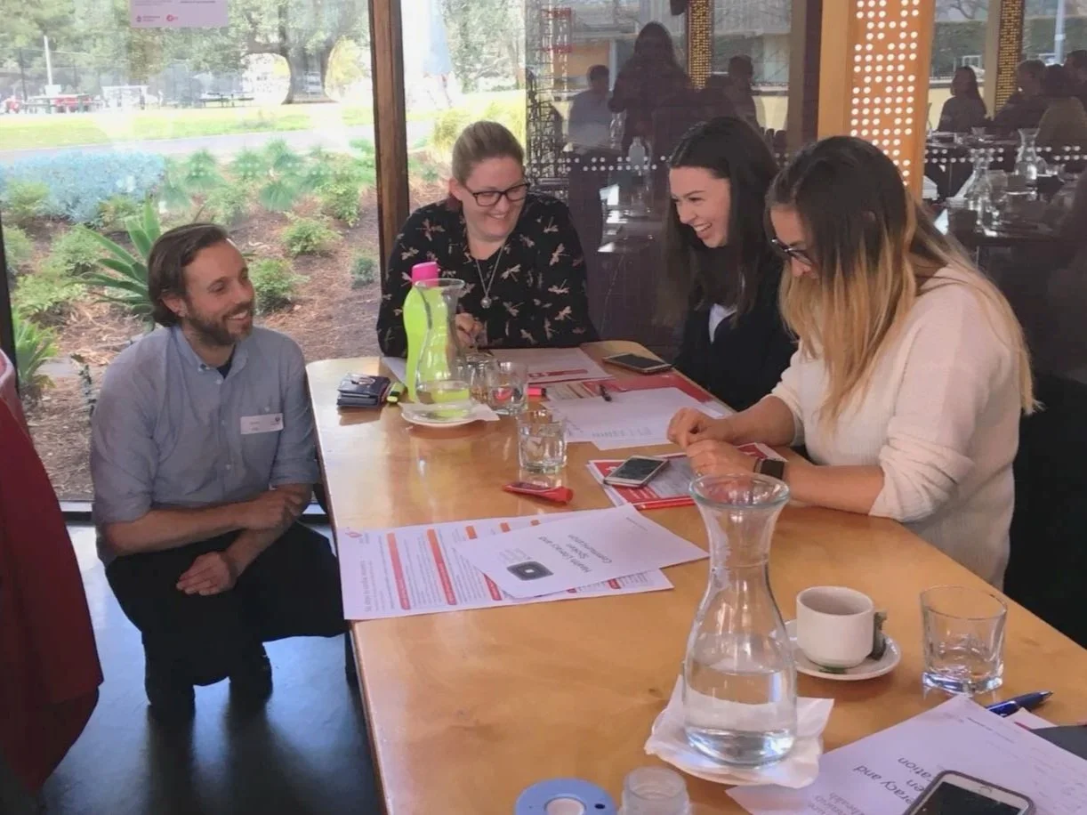 Four people sitting around a table in a restaurant, laughing and engaging in conversation. The table has papers, glasses of water, a water pitcher, and a coffee cup. The background shows large windows with a view outside of greenery.